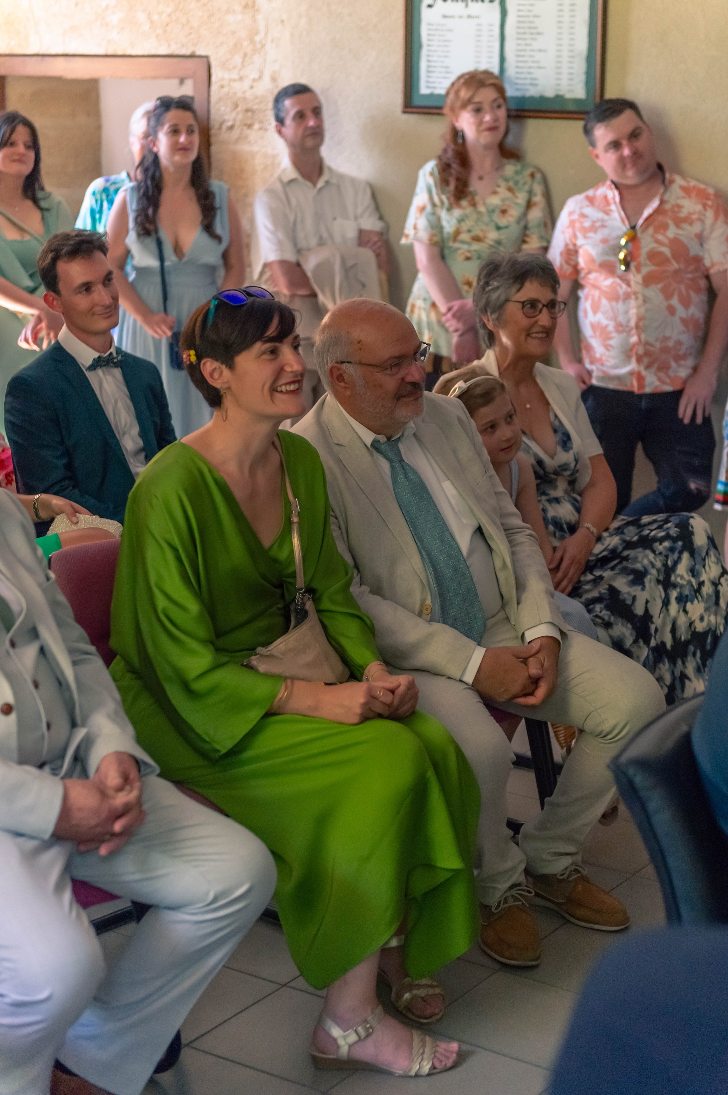 Groupe de personnes assises et debout, assistant à un événement social ou familial dans une pièce avec mur beige et tableau d'affichage en arrière-plan.