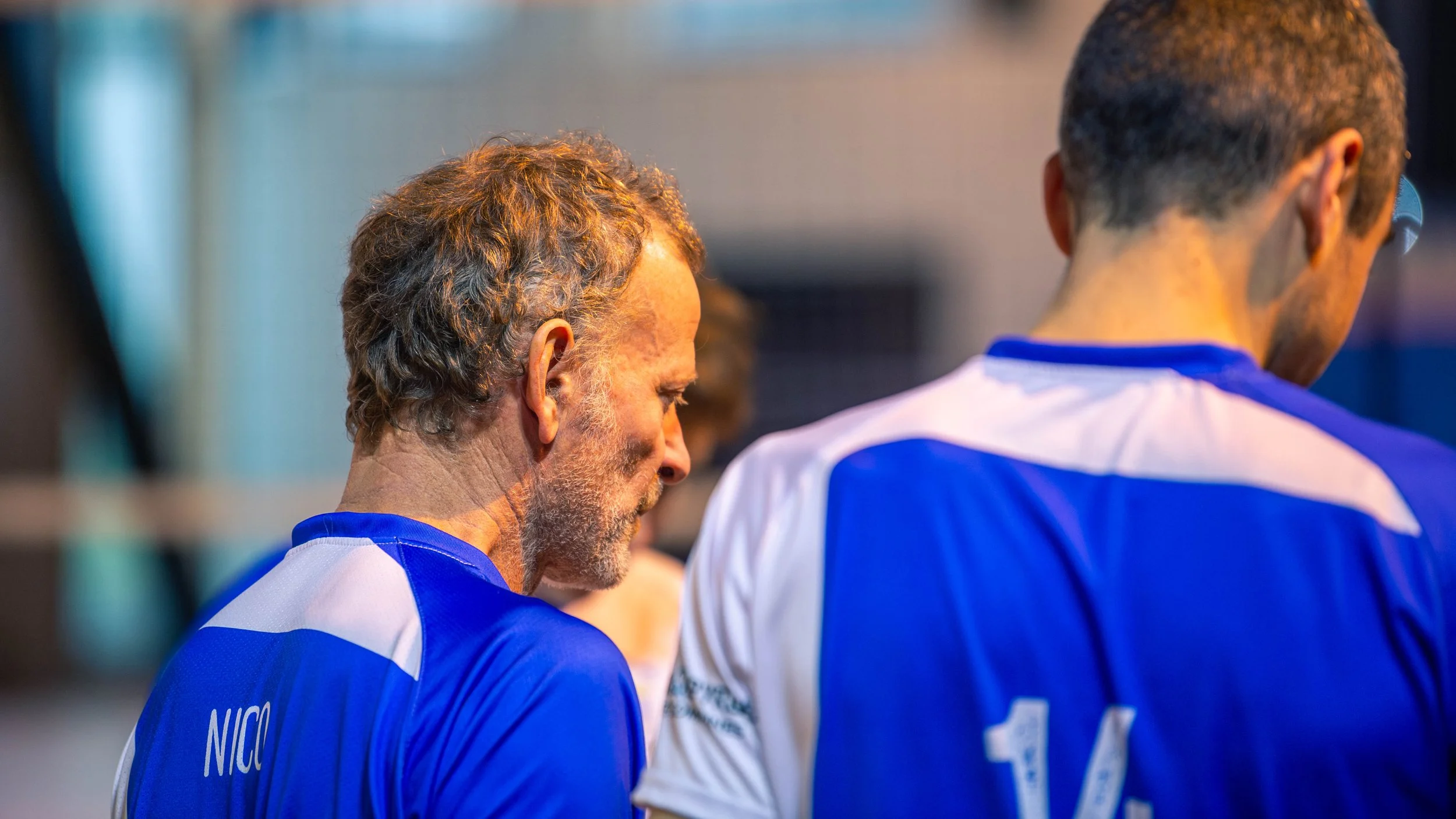 Deux hommes en maillots bleus, vus de dos, probablement des athlètes, se tiennent en silence ou en méditation dans une salle.