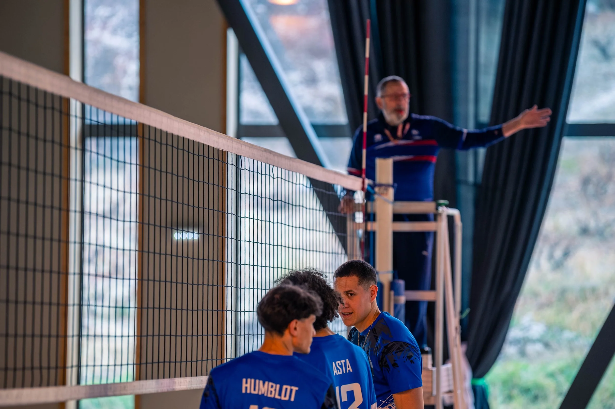 Trois joueurs de volleyball discutent au bord du terrain, un entraîneur en arrière-plan donne des instructions lors d'un match en intérieur.