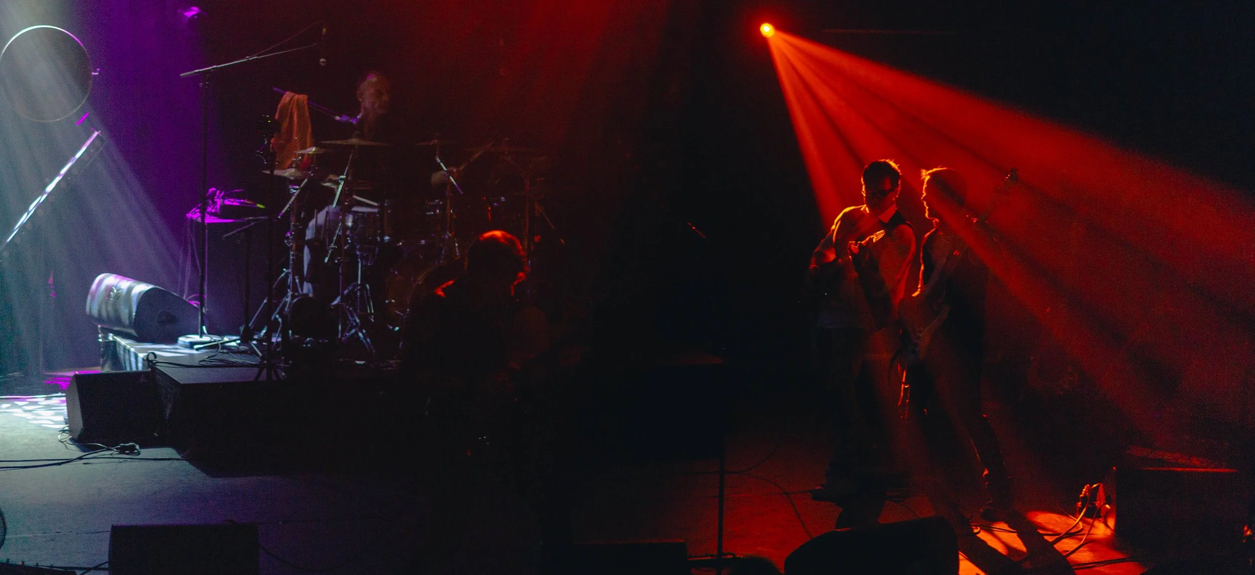 Scène de concert avec trois musiciens, deux à gauche jouant de la guitare sous un éclairage rouge, et un batteur en arrière-plan sous des lumières violettes et blanches.