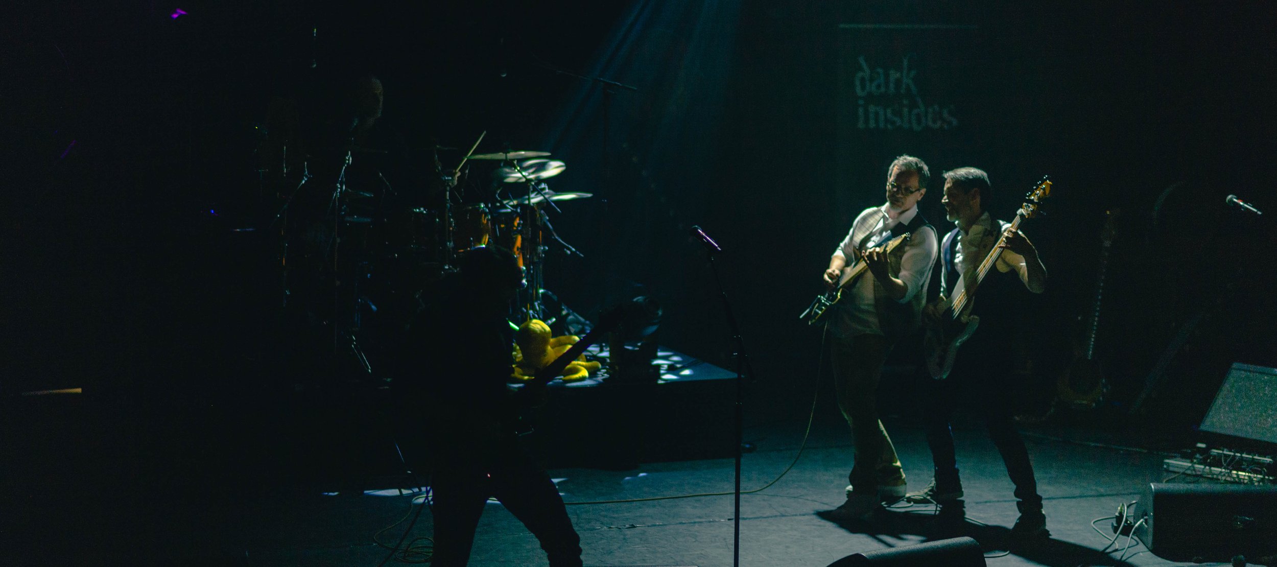 Groupe de musiciens jouant en concert dans une scène sombre, un des musiciens à la guitare et un autre à la basse, avec un fond où il est écrit 'dark insidous'.