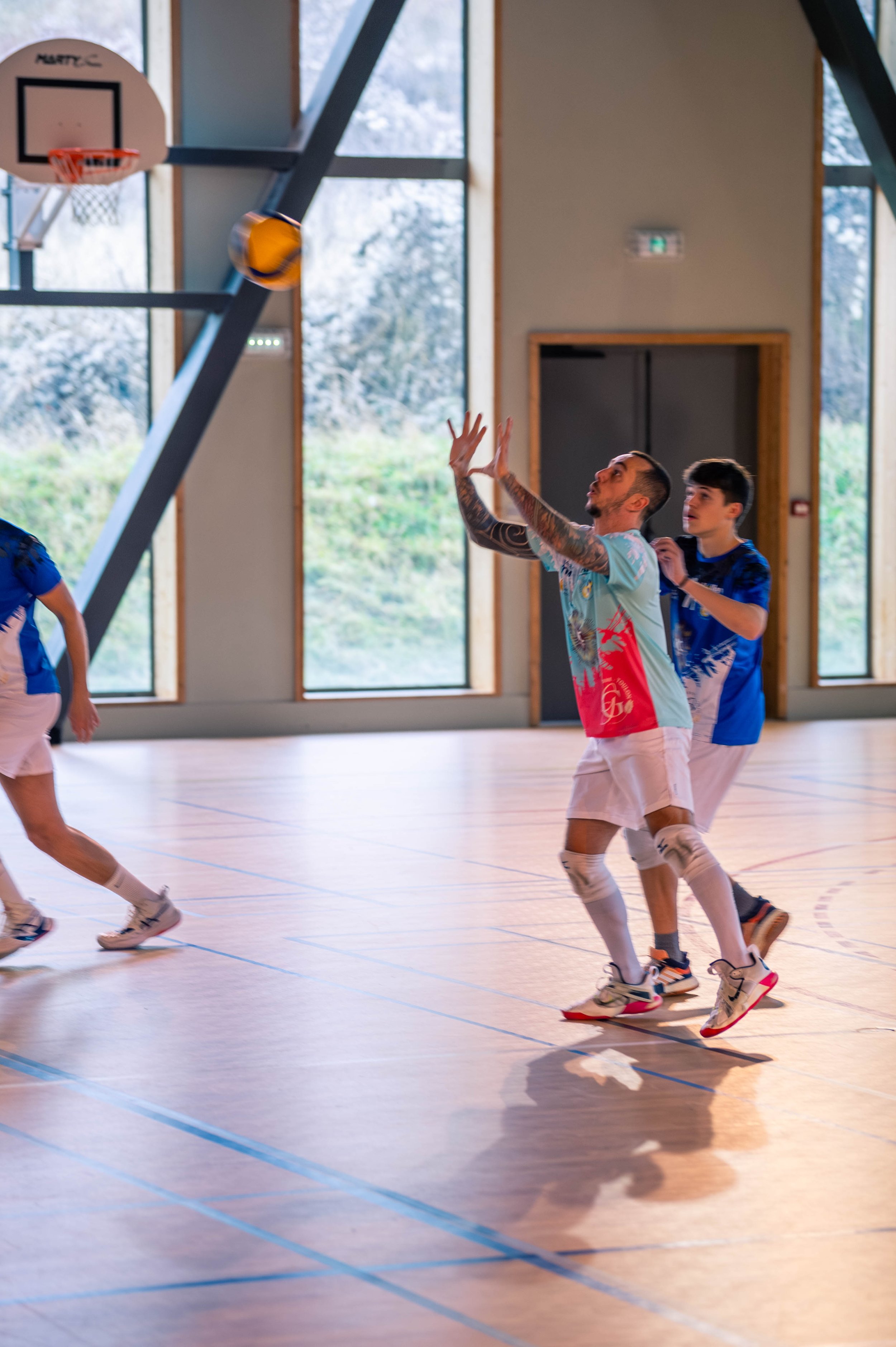 Des joueurs de basketball en train de jouer dans une salle de sport intérieure, avec un ballon en l'air et une table de basketball suspendue au plafond.