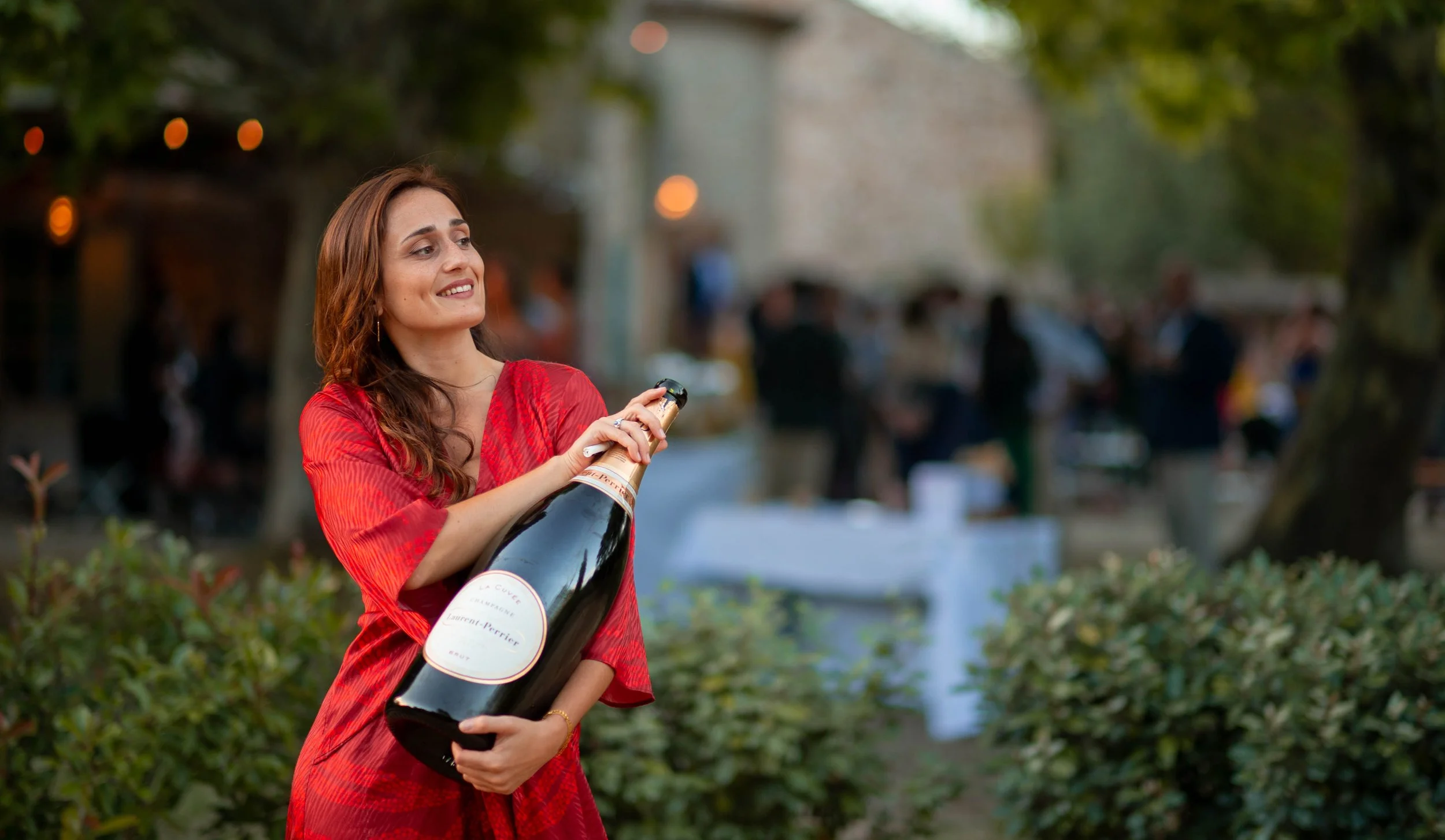 Femme en robe rouge tenant une grande bouteille de champagne lors d'un événement en plein air.