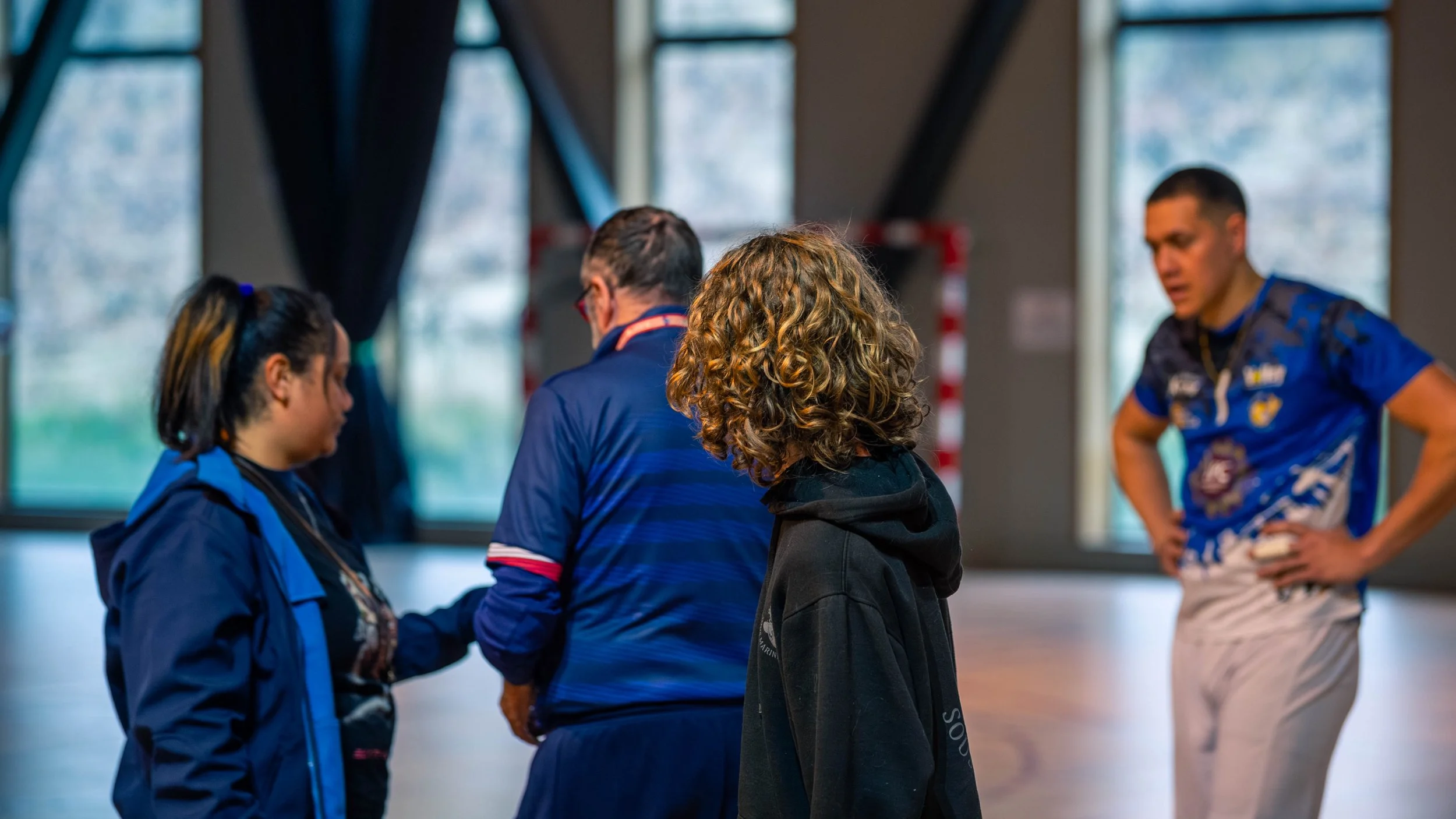 Quatre personnes en intérieur, deux femmes et deux hommes, semblent discuter après une séance ou un match de sport, portant des vêtements de sport.