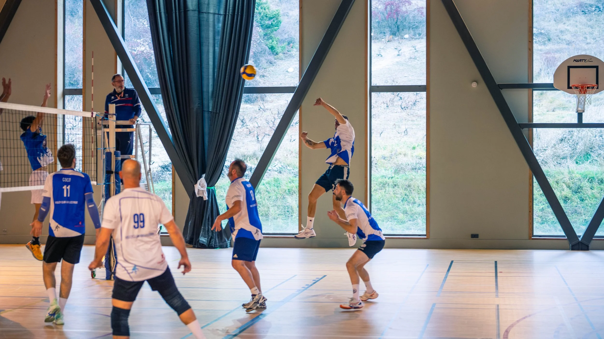 Une partie de volley-ball en intérieur, des joueurs en action, un spectateur en arrière-plan, et un terrain de sport avec un mur en verre et un basketball