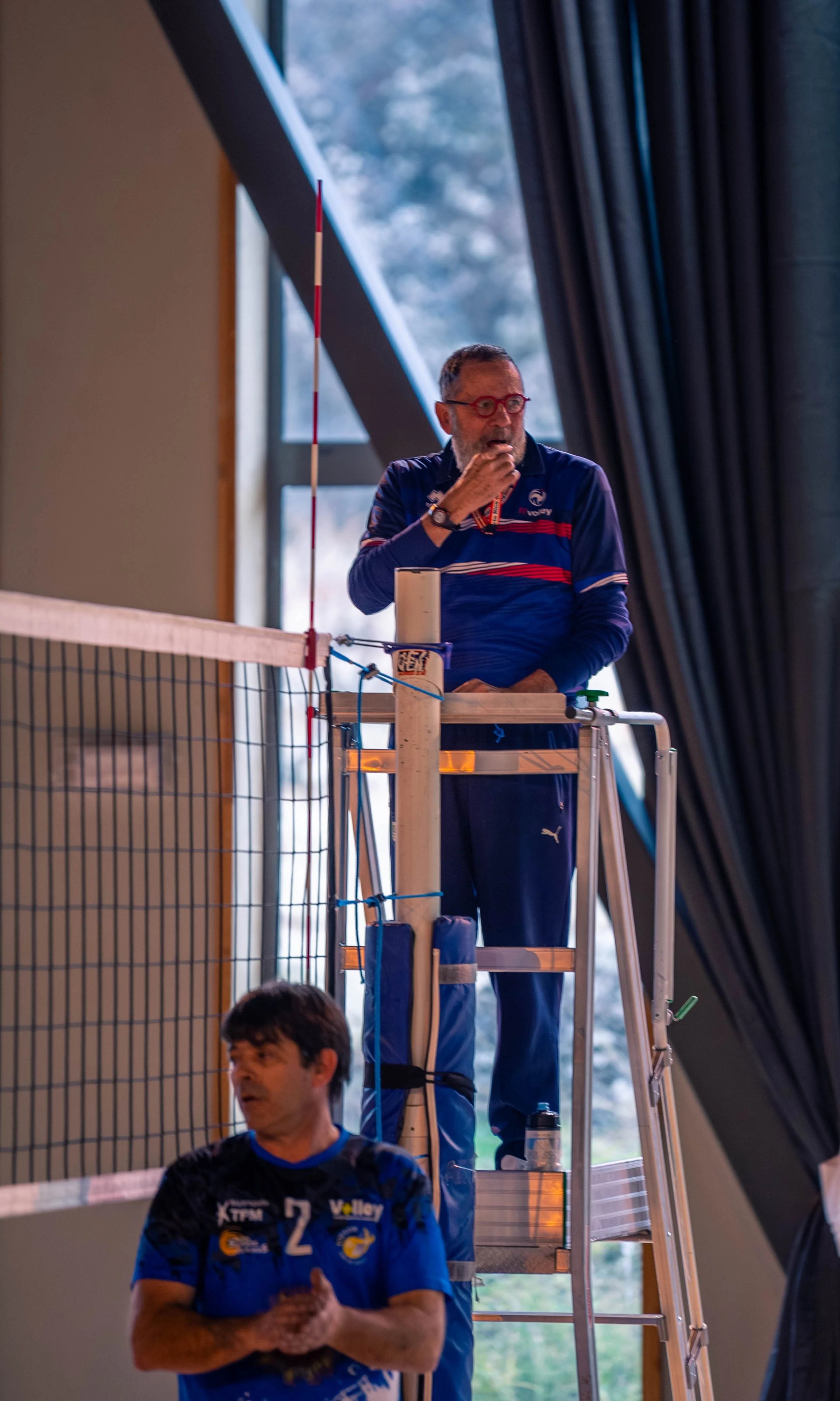 Deux entraîneurs de volleyball en tenue bleue discutent lors d'un match, l'un sur une plateforme en hauteur et l'autre à côté du filet, à l'intérieur d'une salle avec fenêtre et rideaux noirs.