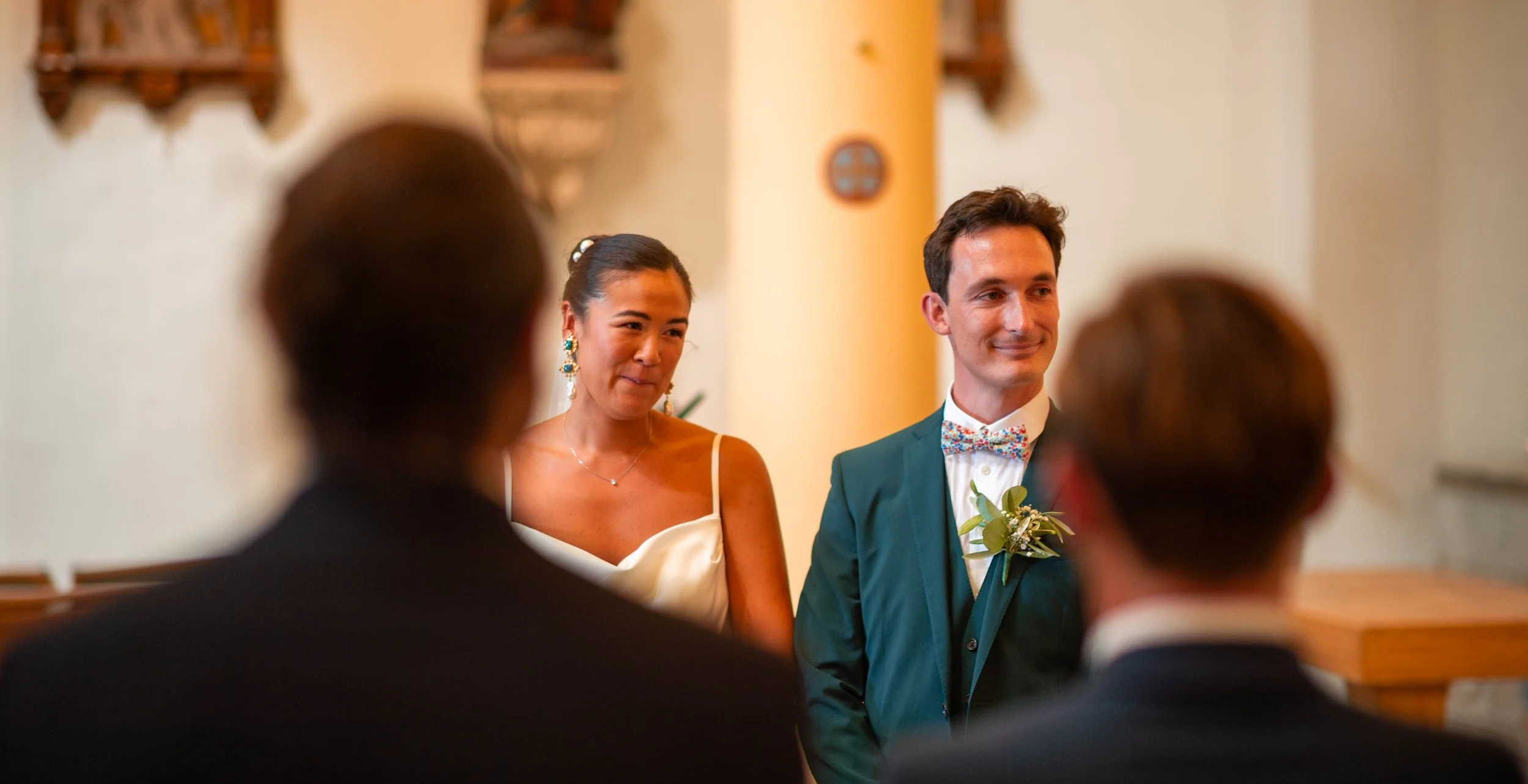 Un couple en costume lors d'une cérémonie de mariage devant deux personnes dont le dos est visible