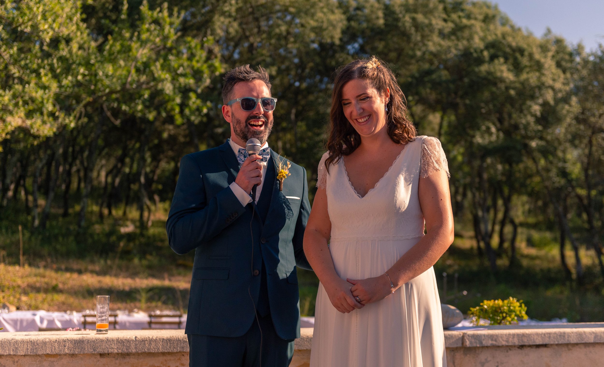 Un homme en costume bleu, portant des lunettes de soleil et une lavallière, parle dans un microphone lors d'une cérémonie en plein air. Une femme en robe blanche l'écoute, souriante, dans un décor de nature avec des arbres en arrière-plan.