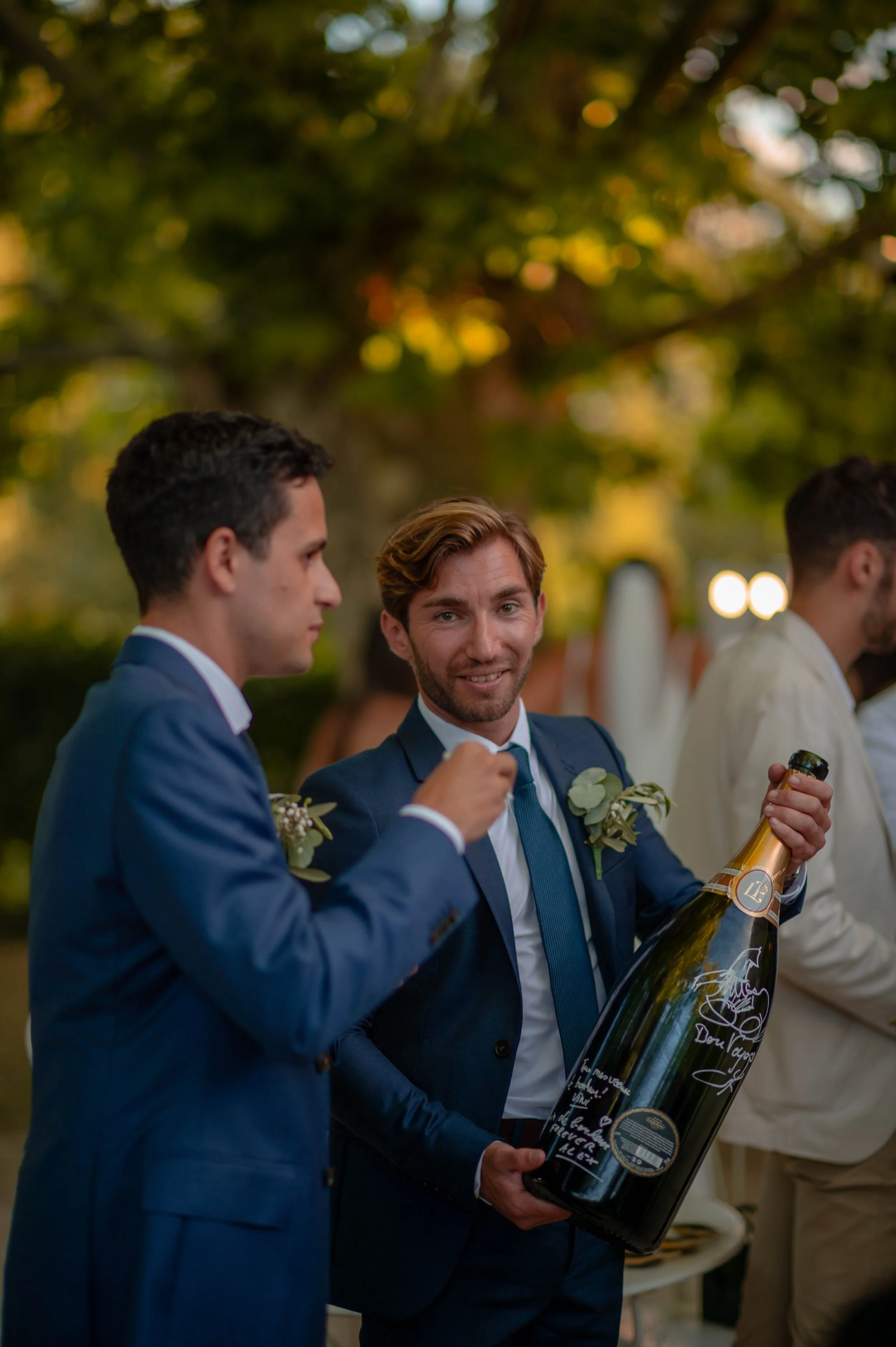 Deux hommes en costume lors d'un mariage ou d'une célébration, l'un d'eux tient une grande bouteille de champagne et les deux portent des boutonnières, avec un décor naturel en arrière-plan.