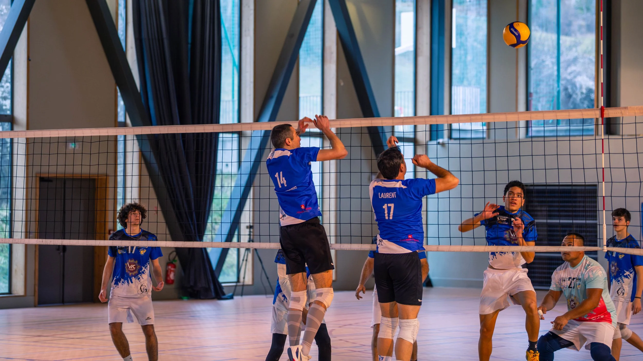 Une équipe de volley-ball en action dans un gymnase intérieur, avec plusieurs joueurs en bleu et un en blanc, des fenêtres en arrière-plan et un ballon orange et bleu en vol