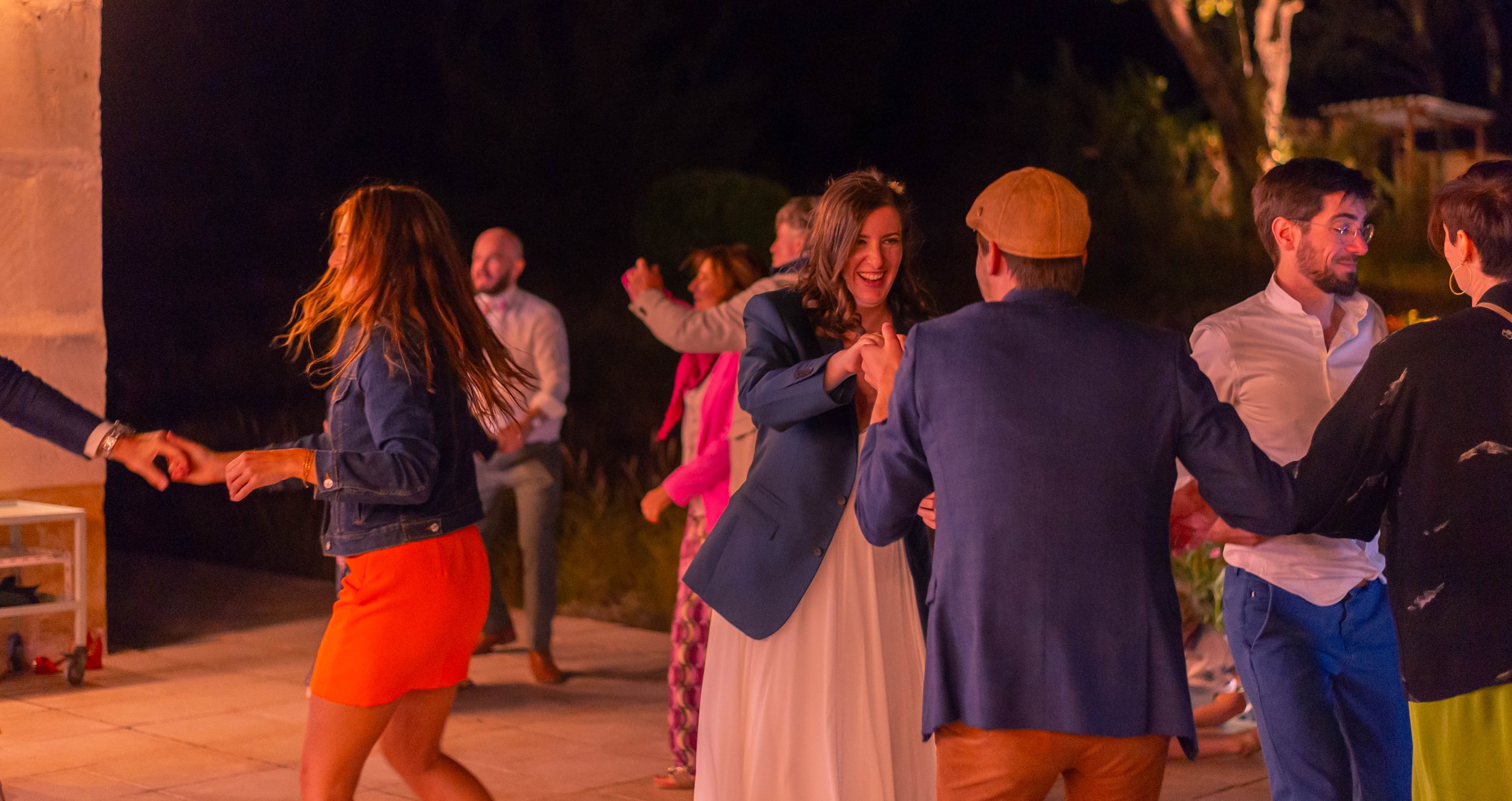 Groupe de personnes dansant et souriant lors d'une soirée en plein air la nuit, éclairée par des lumières chaleureuses.
