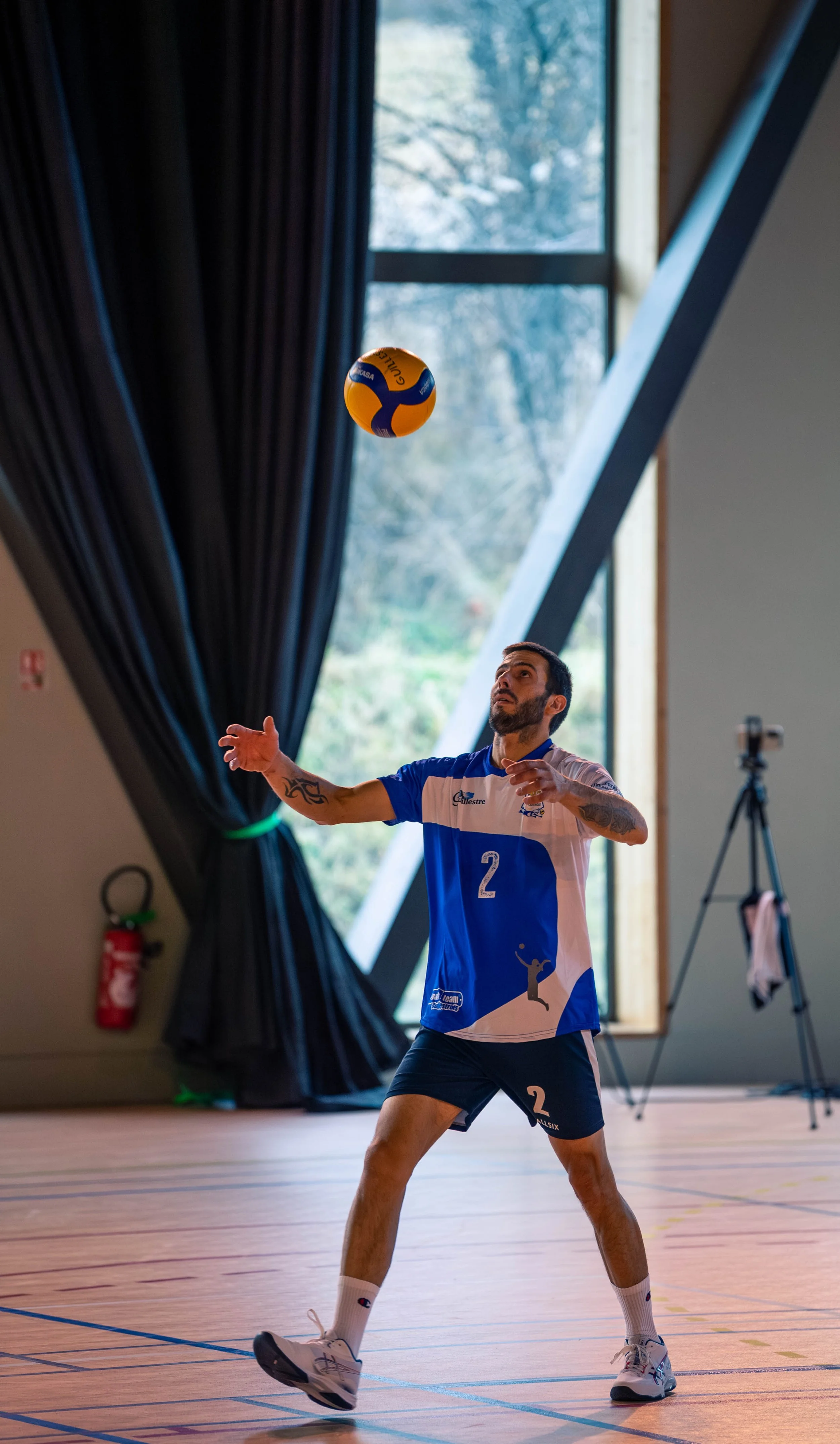 Un homme joue au volleyball intérieur, portant un maillot bleu et blanc, sautant pour attraper une balle orange et bleue en salle, avec un fond de fenêtres laissant entrer la lumière naturelle.
