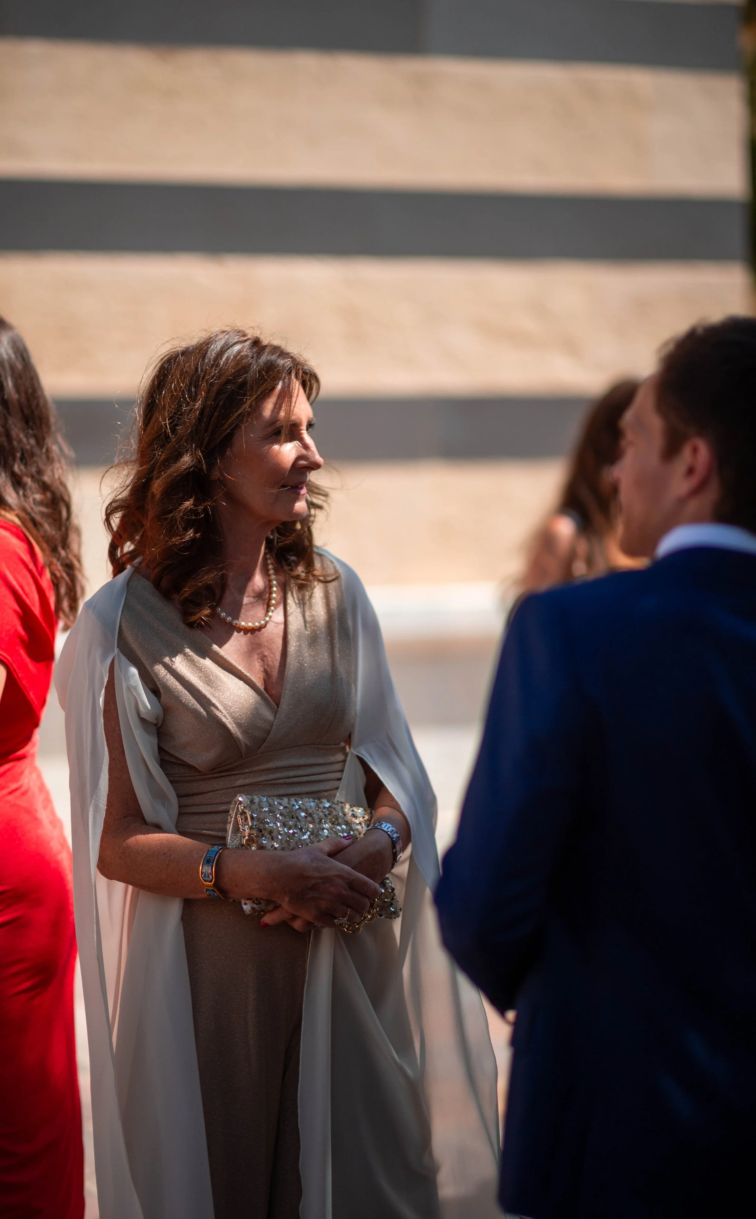 Femme âgée portant une robe beige, un collier de perles, tenant un sac à main orné de bijoux, discutant avec un homme en costume bleu, lors d'un événement social.