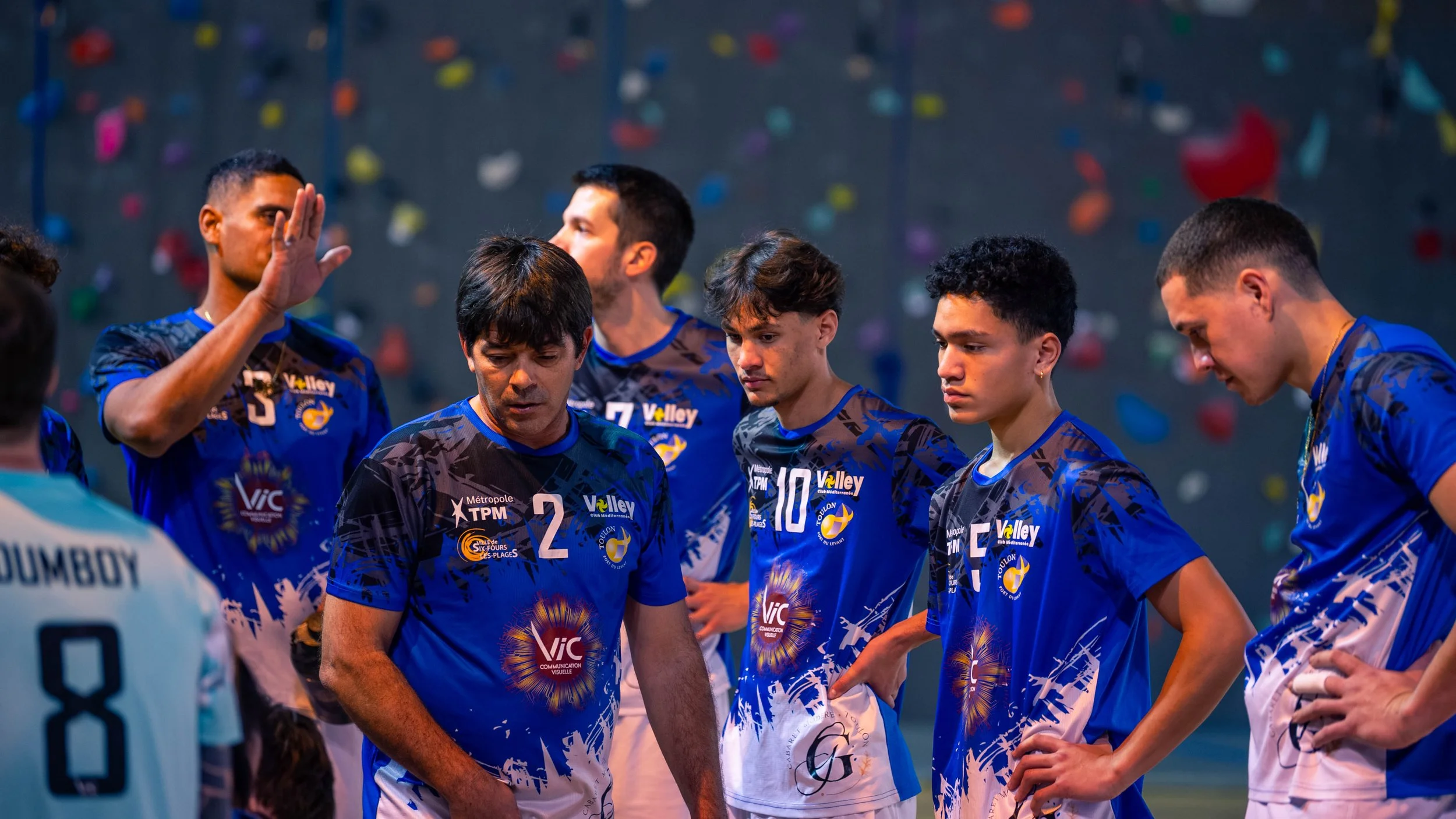 Une équipe de volleyball en tenue bleue écoute probablement leur entraîneur lors d'une discussion pendant un match ou une entraînement, en arrière-plan un mur avec une structure d'escalade.