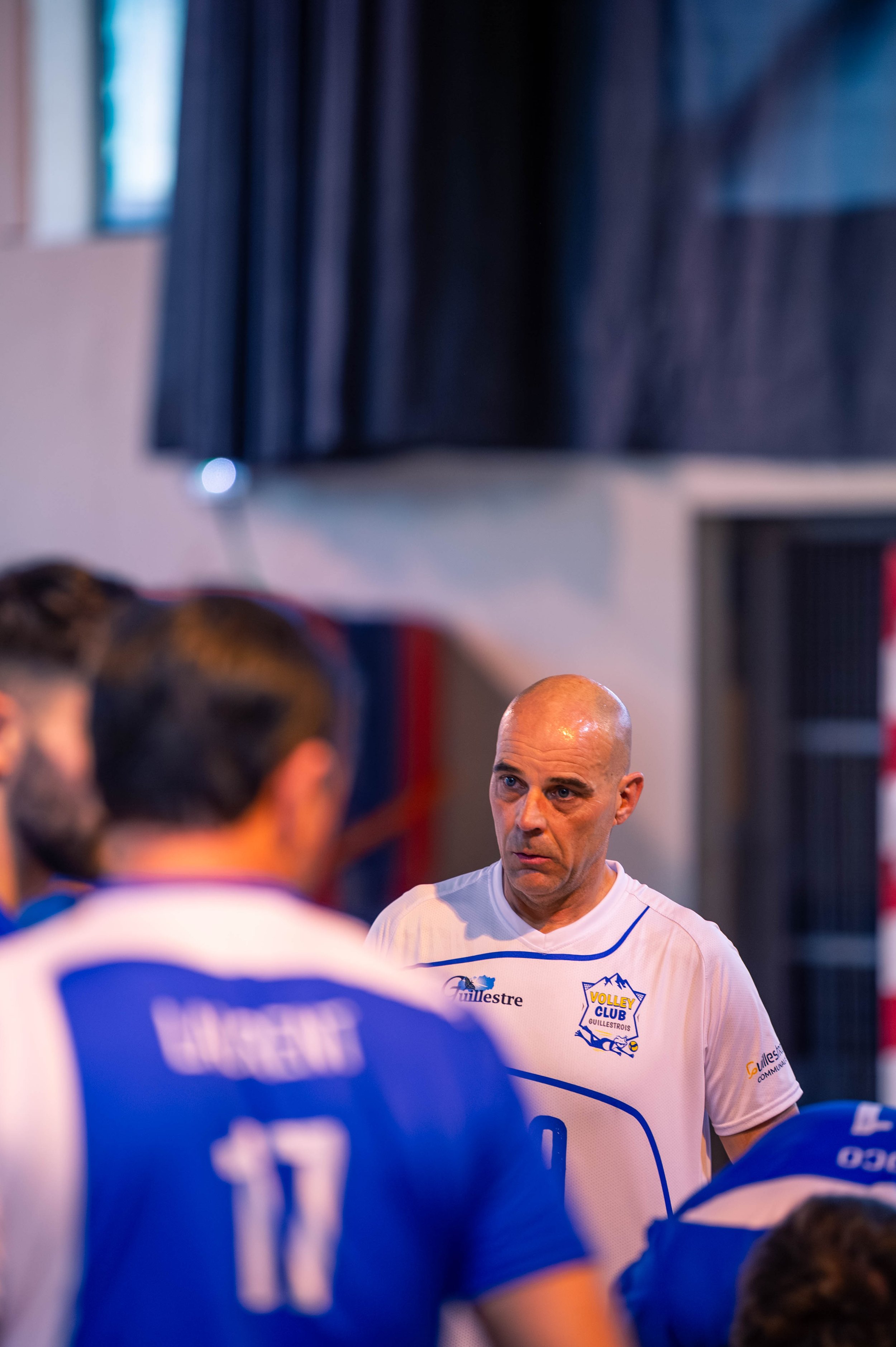 Un entraîneur de volley-ball parle à ses joueurs lors d'une séance d'entraînement ou d'une pause d'équipe, tous portant des maillots bleus et blancs.