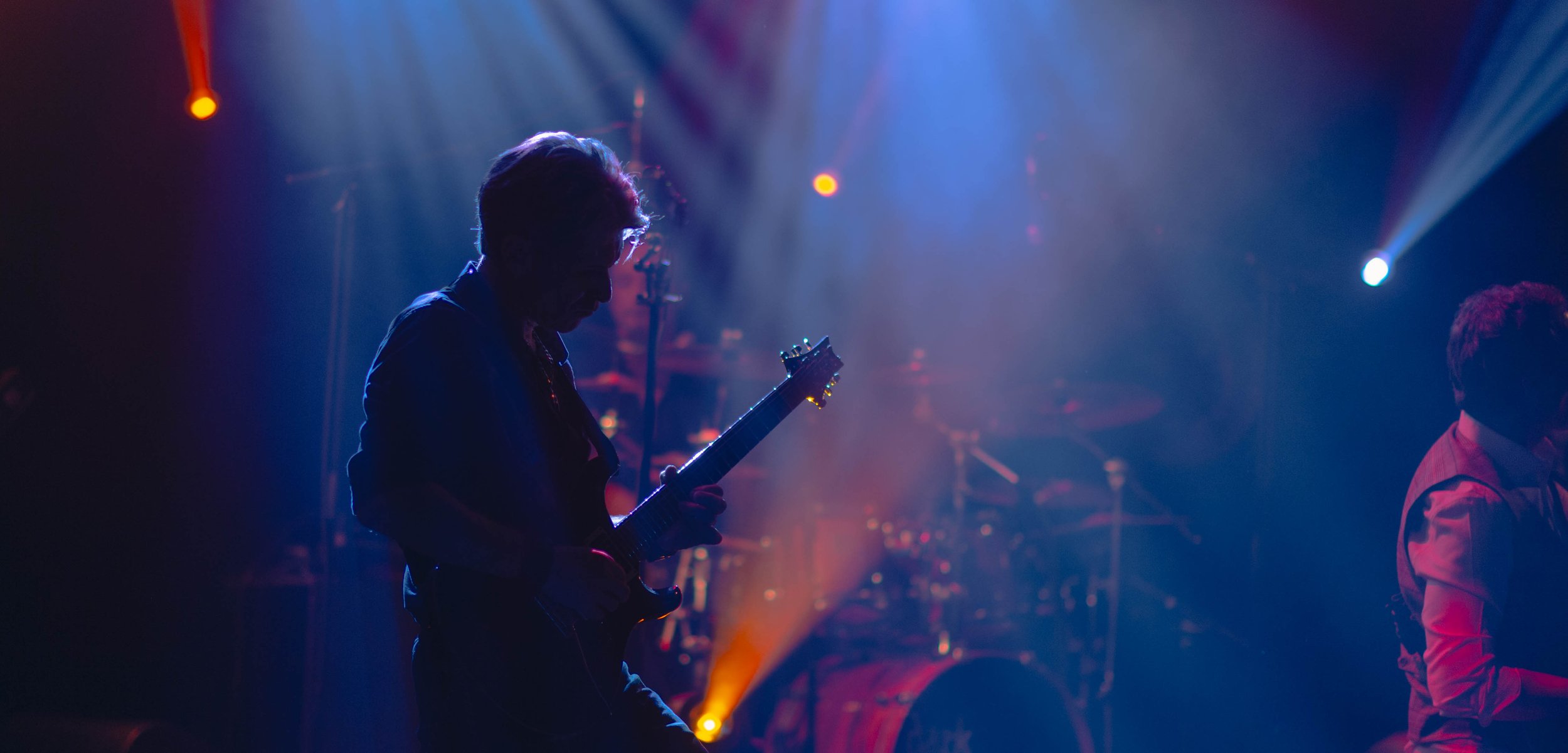 Musiciens jouant de la guitare et du piano sur scène lors d'un concert, sous un éclairage coloré avec des effets de lumière.
