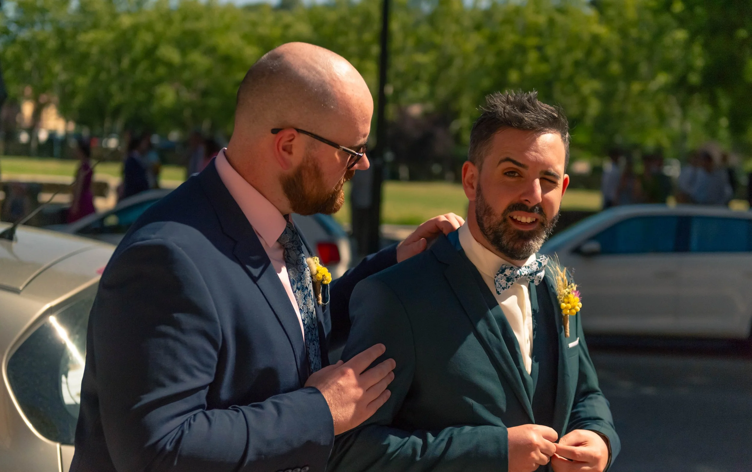 Deux hommes habillés en costume, l'un semble consoler l'autre, dans un lieu extérieur avec des voitures en arrière-plan.