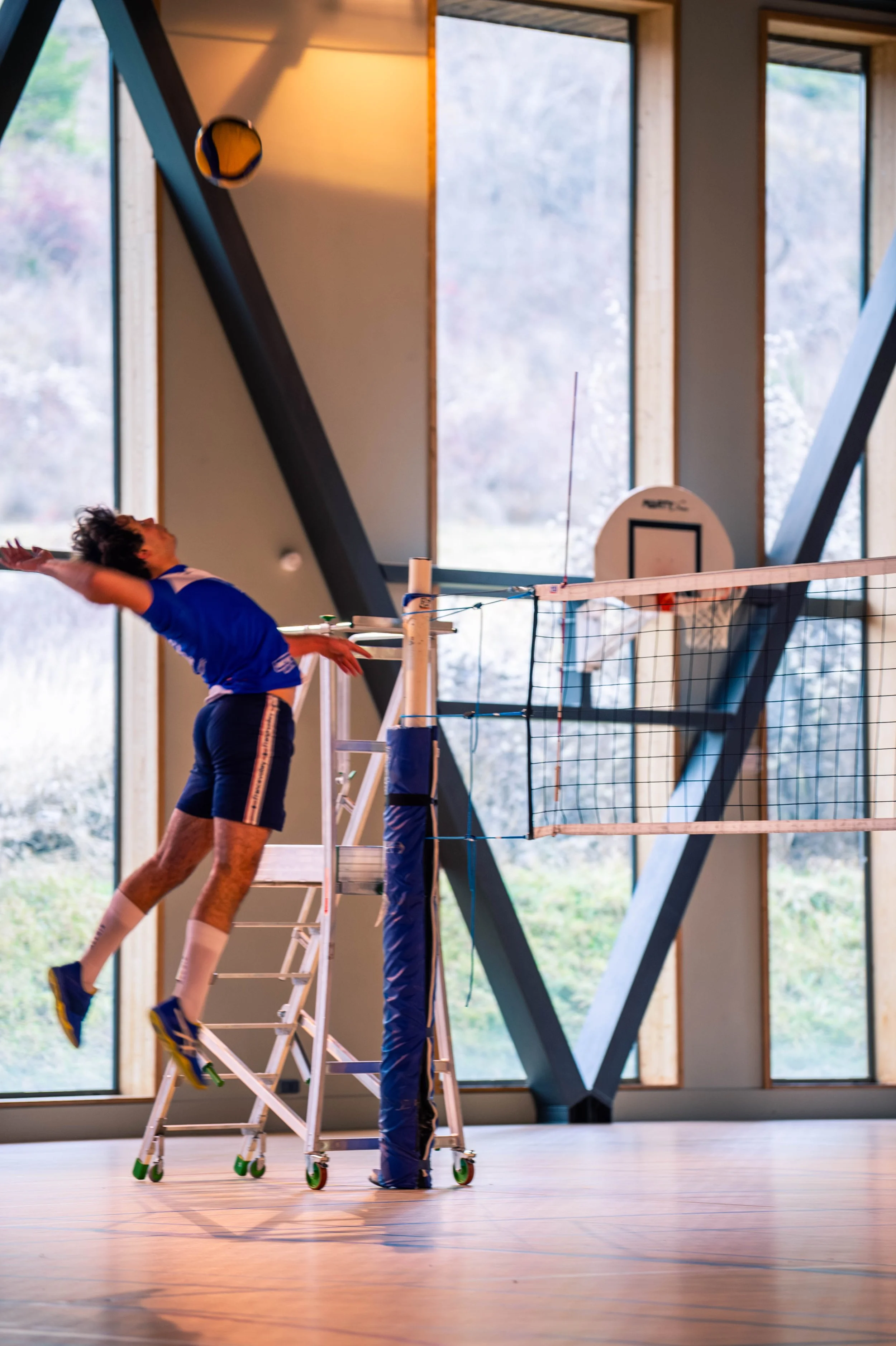 Un joueur de volley-ball est en train de sauter pour frapper le ballon dans une salle intérieure avec de grandes fenêtres.