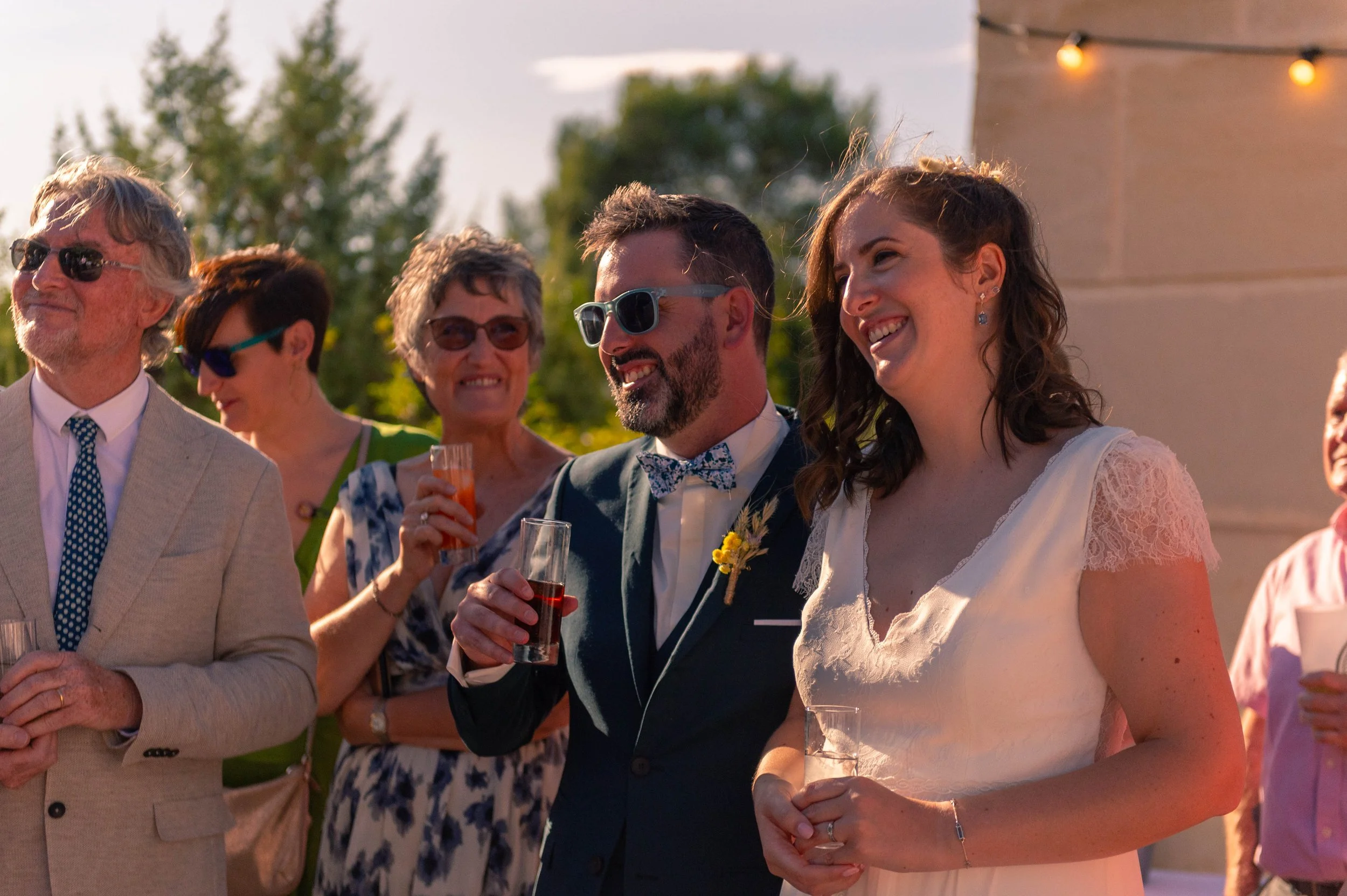 Groupe de personnes lors d'une réception en plein air, célébrant, avec des hommes et des femmes souriants, habillés élégamment, certains portant des lunettes de soleil, tenant des verres de boissons, sous un ciel en soirée avec des lumières suspendue