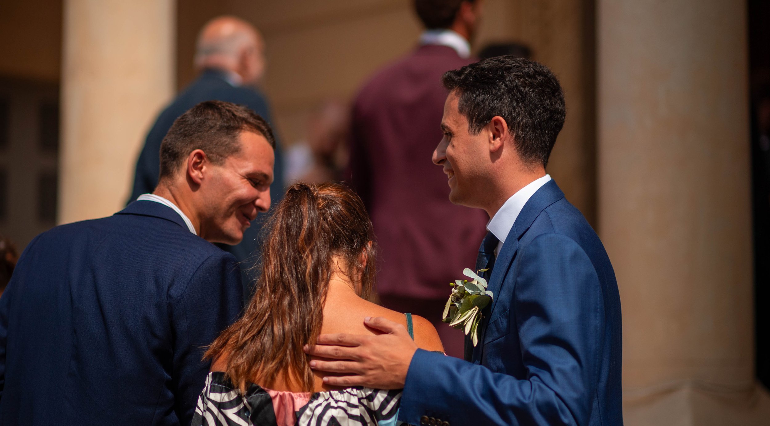 Trois personnes lors d'un mariage, un homme en costume bleu, une femme avec un vêtement d'épaule à motifs, et un autre homme en costume bleu, échangeant un regard et un sourire.