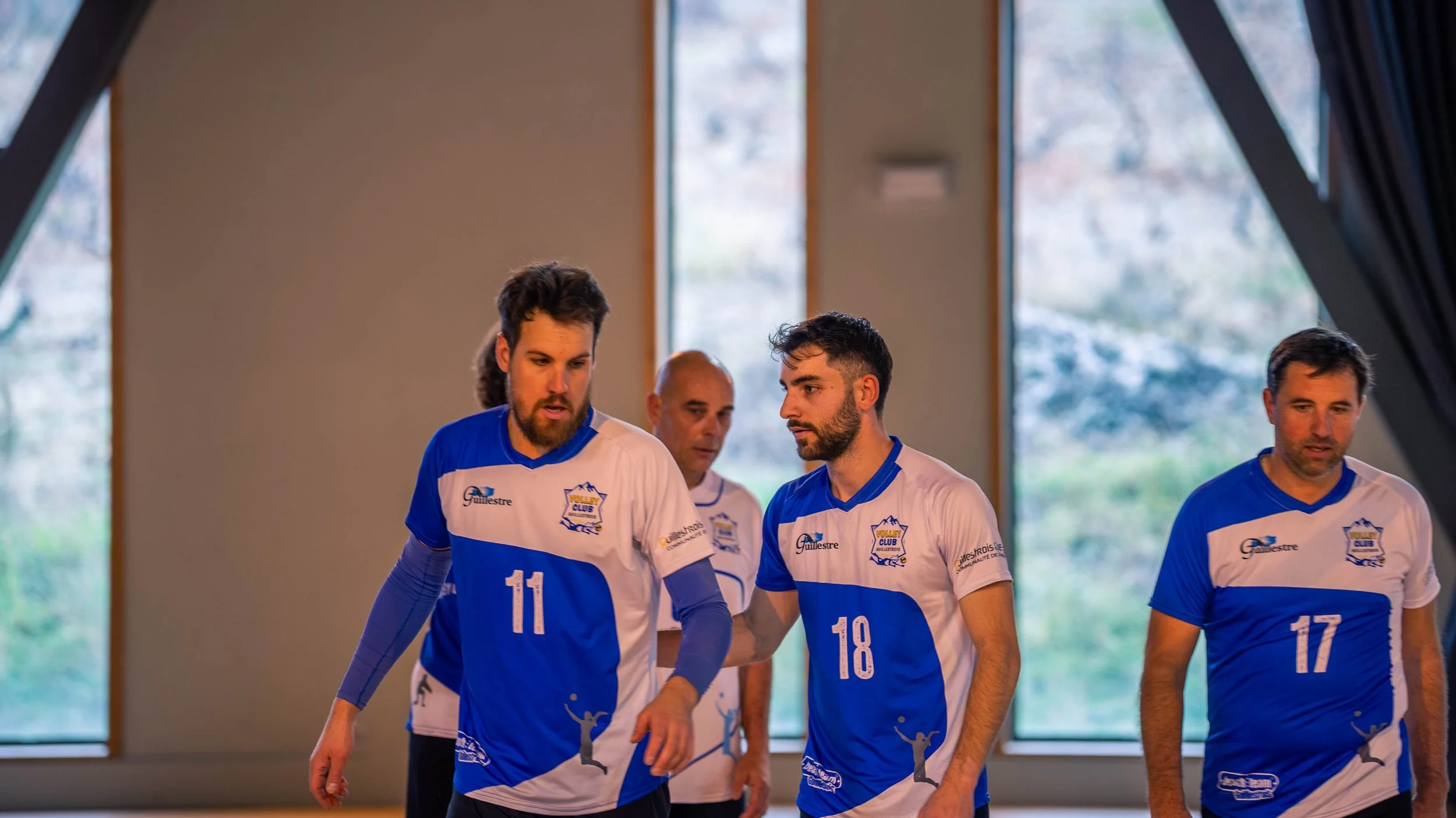 Quatre joueurs de volleyball en tenues bleues et blanches à l'intérieur, avec de grandes fenêtres derrière eux montrant un paysage naturel extérieur.