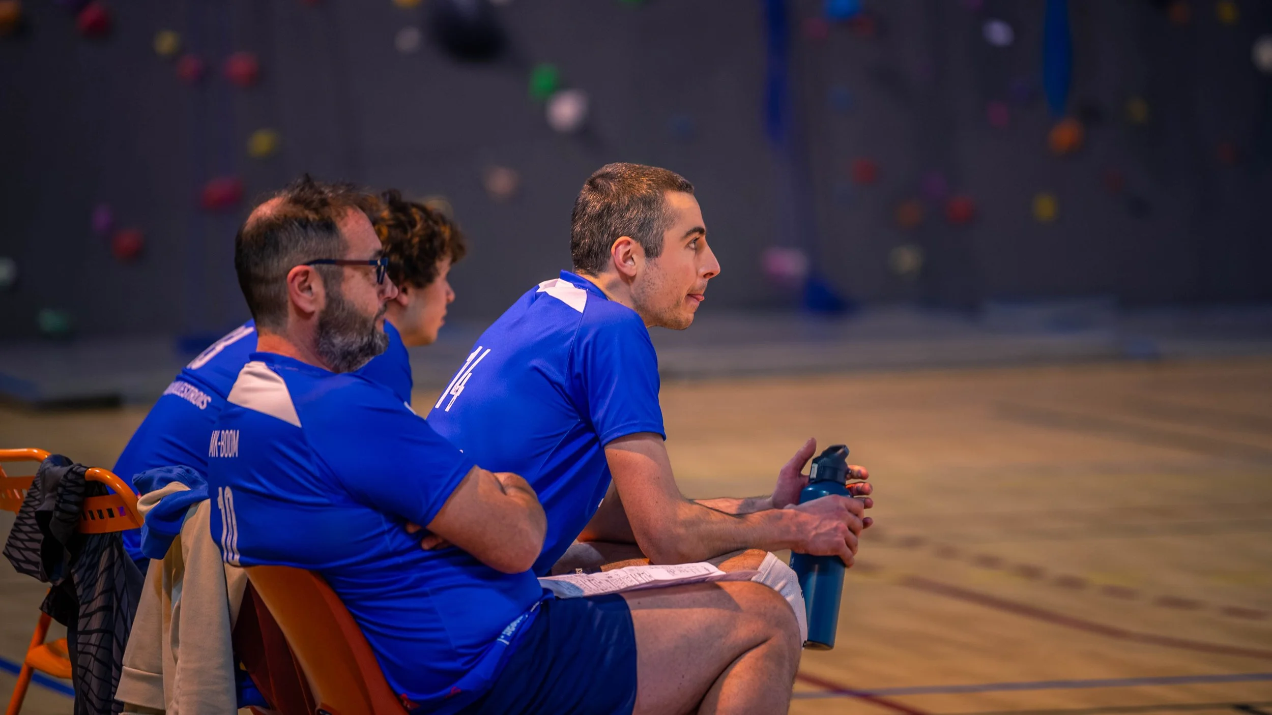 Trois personnes en tenue de sport assises sur des chaises dans une salle de sport ou gymnase, regardant un mur d'escalade en arrière-plan.