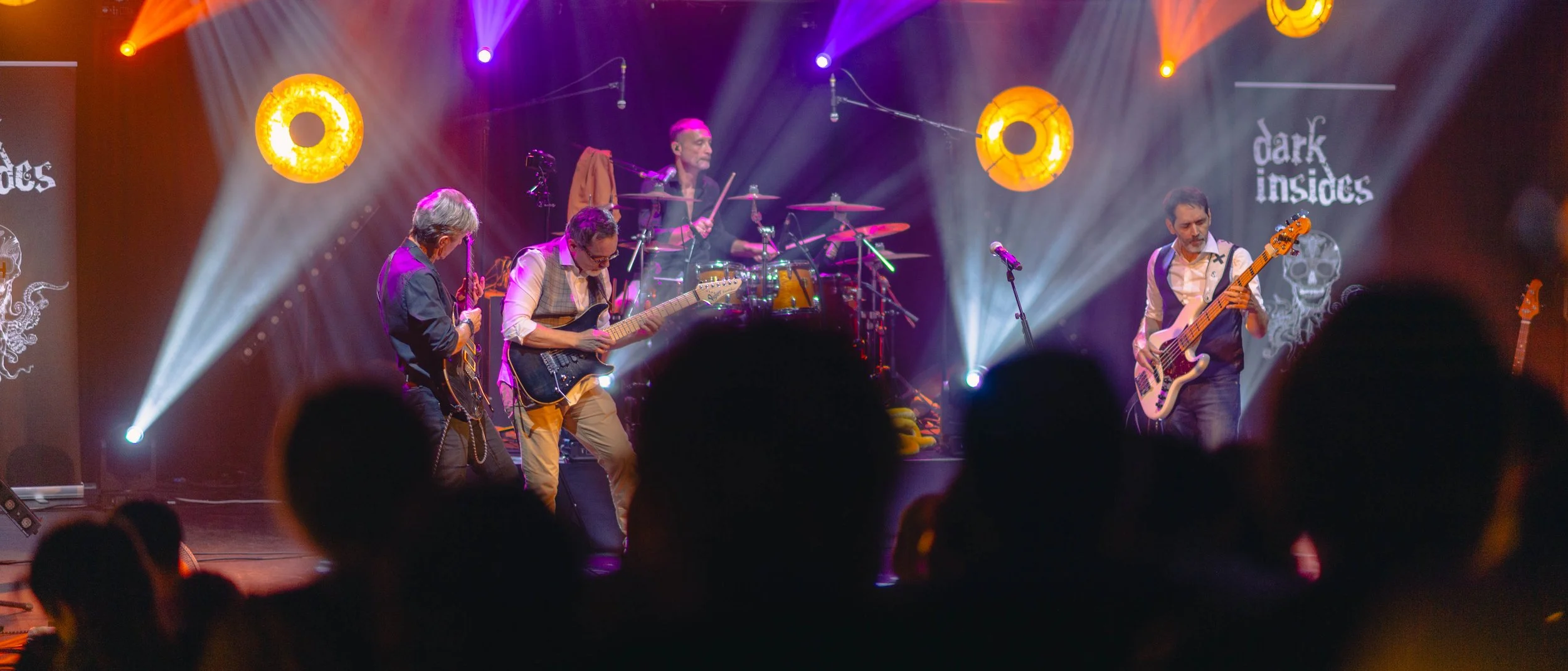 Un groupe de musiciens joue sur scène lors d'un concert, avec un éclairage coloré et un écran en arrière-plan affichant le nom du groupe 'dark insides'.