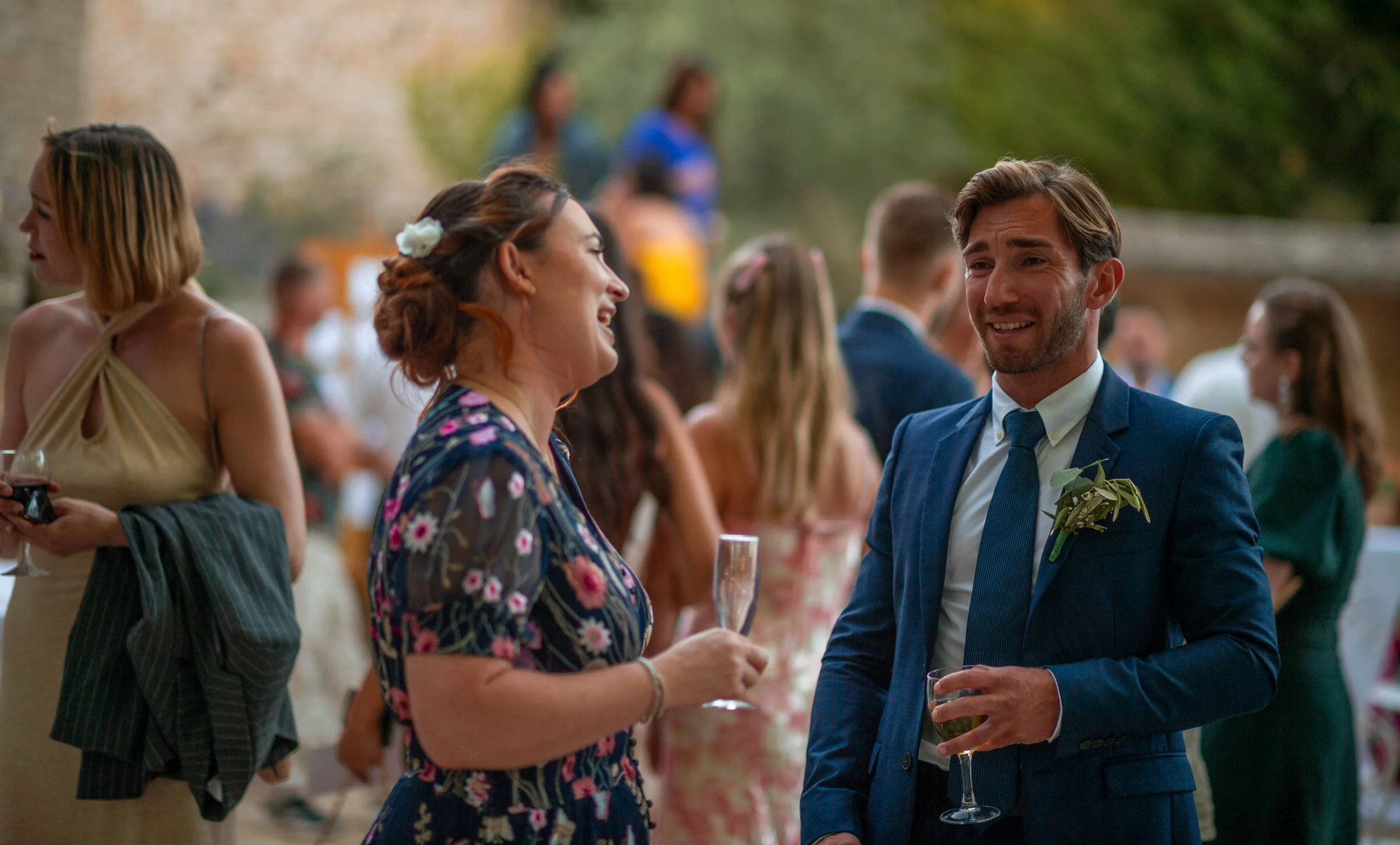 Groupe de personnes lors d'une célébration extérieure, avec deux personnes au premier plan échangeant un sourire et entretenant une conversation, tenant des verres de vin ou de champagne, en vêtements élégants.