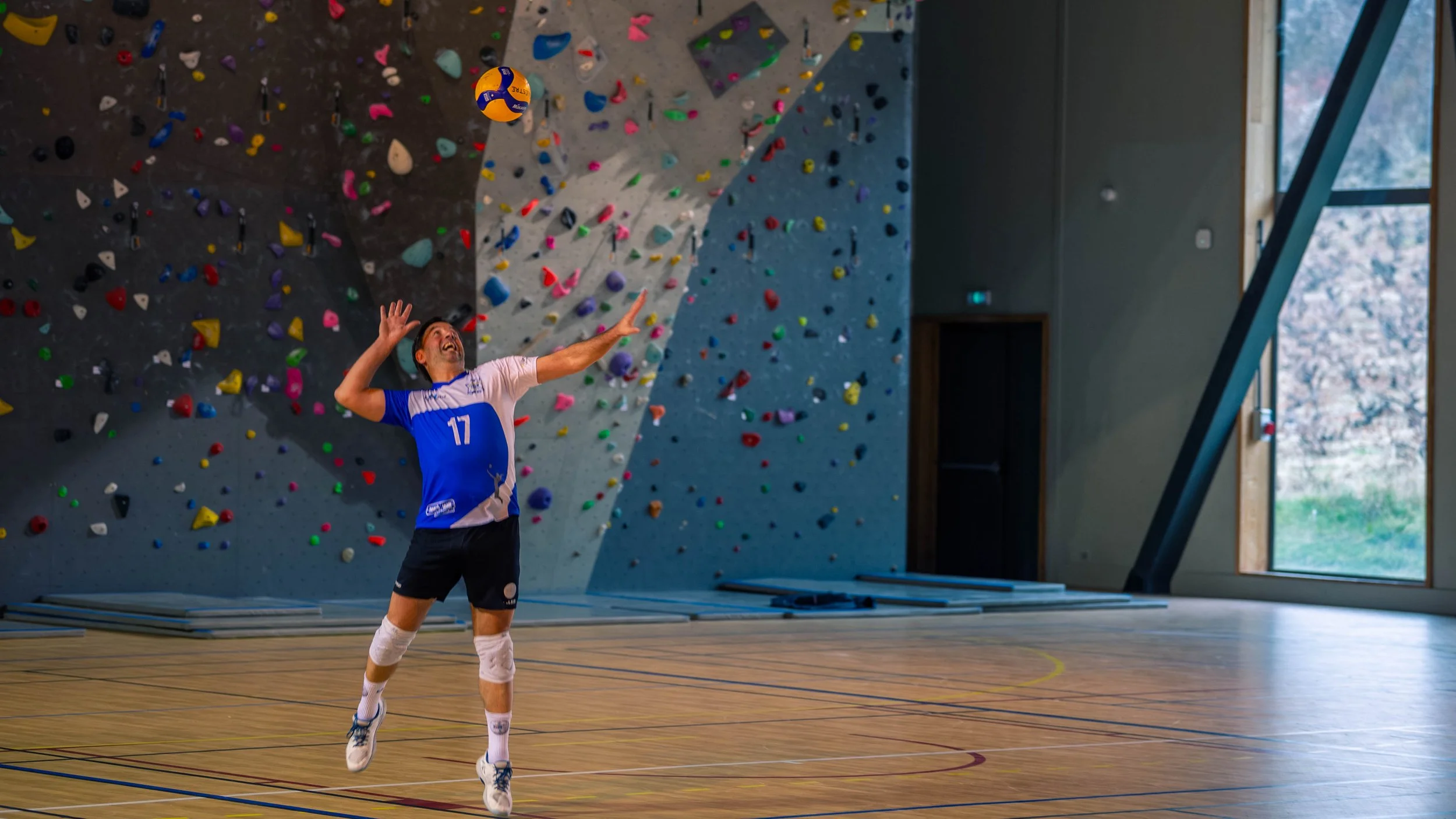 Un joueur de volley-ball en plein saut dans un gymnase intérieur, prêt à frapper le ballon, avec un mur d'escalade en arrière-plan.