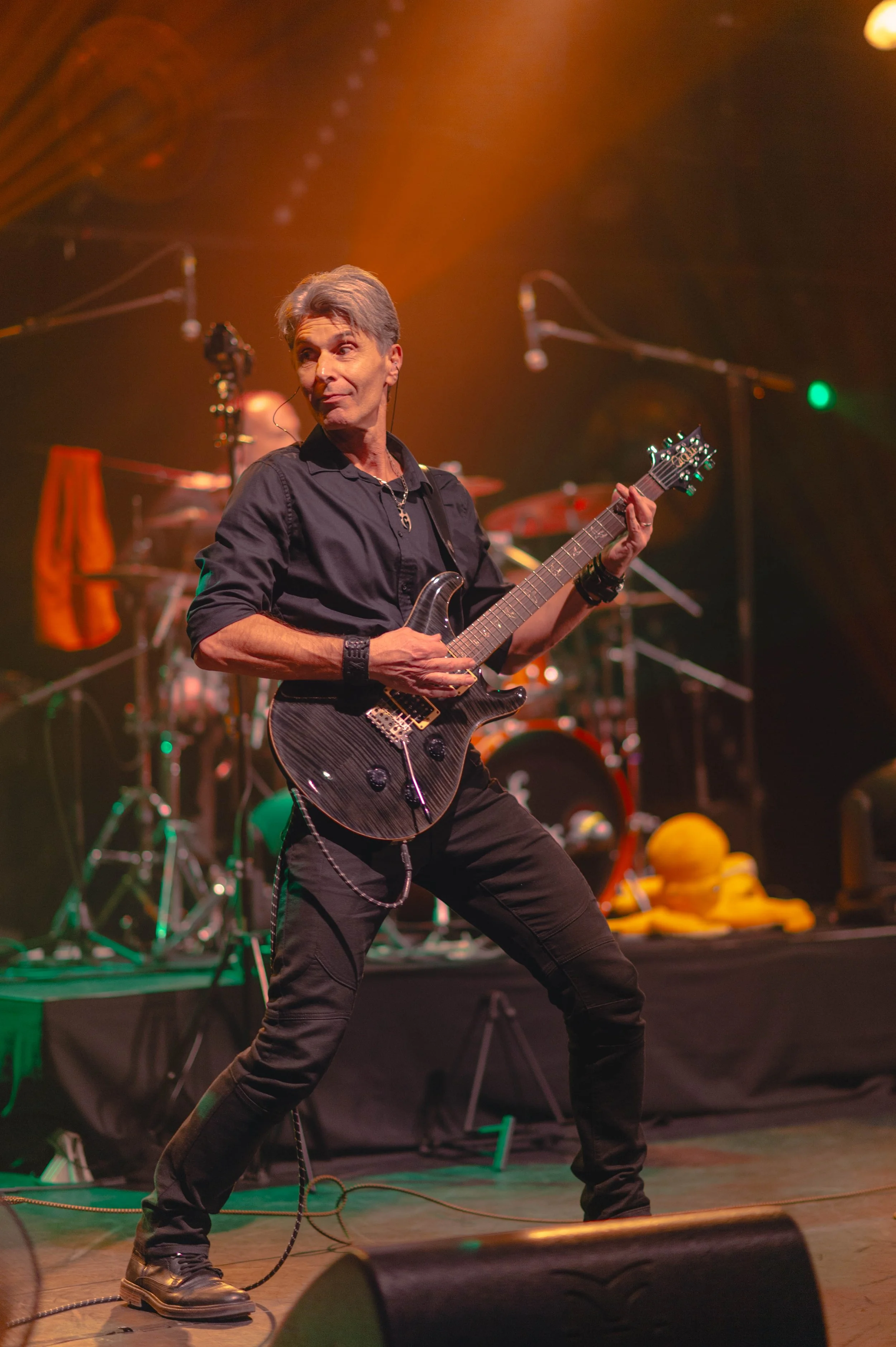 Musicien jouant de la guitare en concert, avec batterie en arrière-plan, sous un éclairage coloré