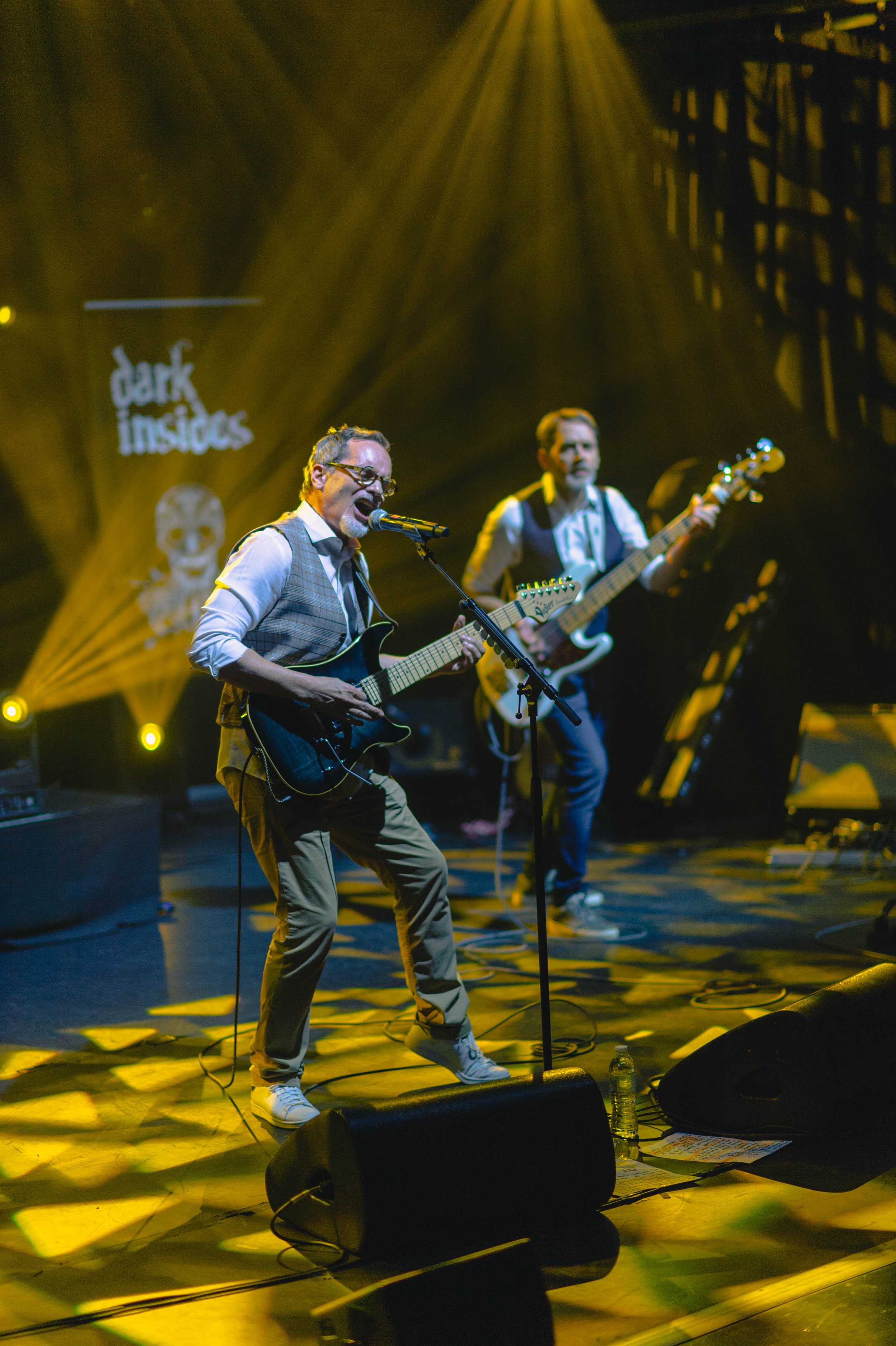 Deux musiciens jouent de la guitare électrique lors d'un concert sur scène, avec des lumières jaunes et un fonds affichant 'dark insides'.