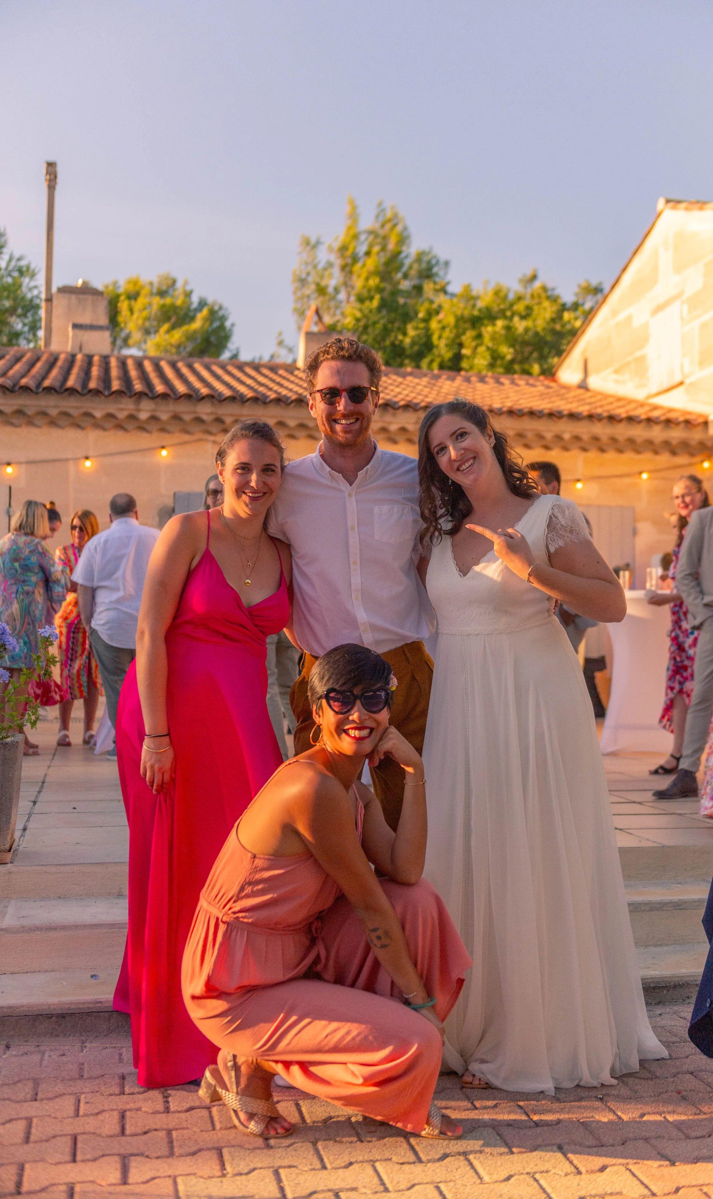 Groupe de quatre personnes souriantes lors d'une fête en plein air, deux femmes en robes roses et blanches, un homme en chemise blanche et pantalon marron, une femme en robe pêche avec des lunettes noires. Atmosphère chaleureuse avec des lumières et 