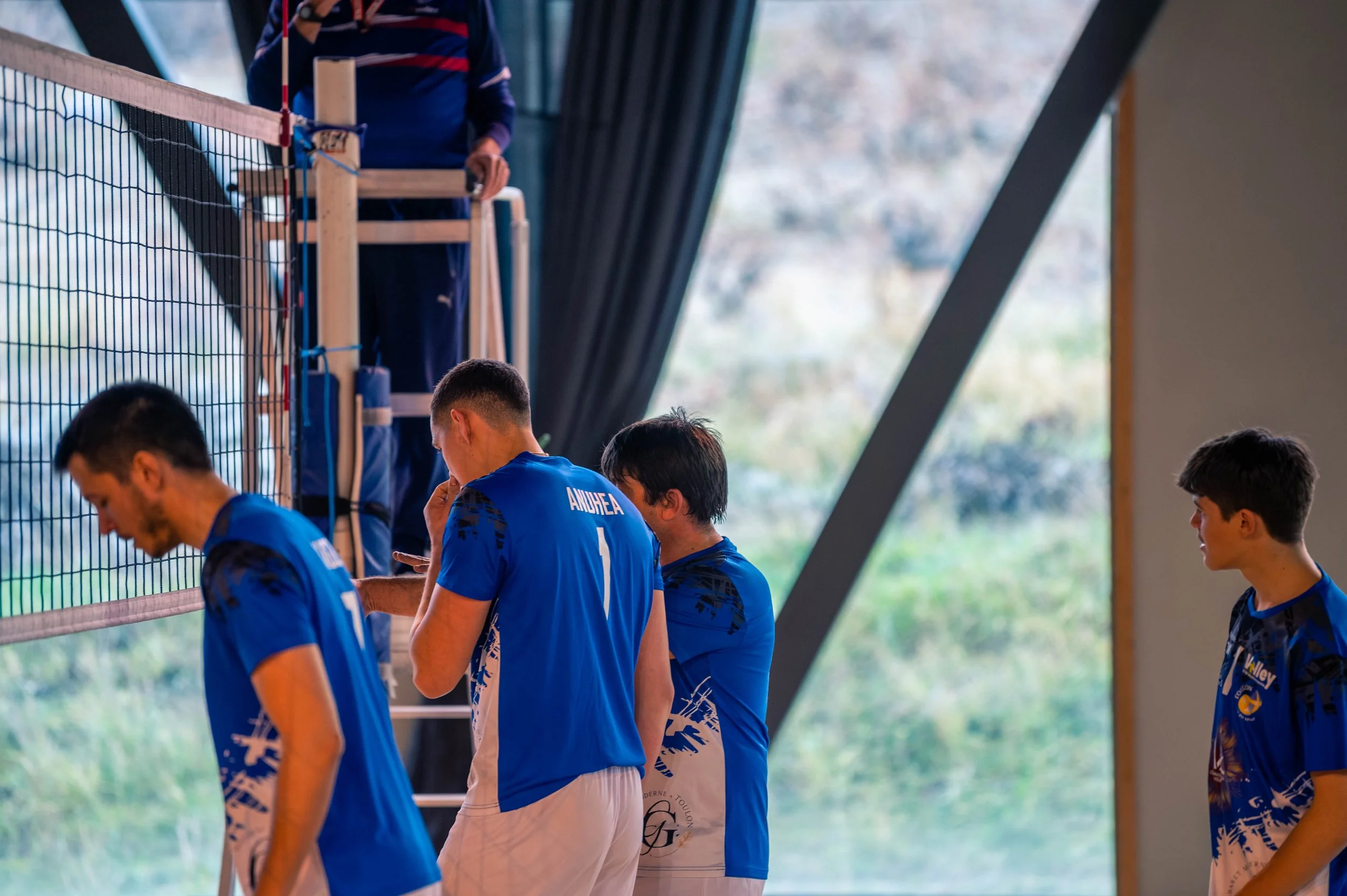 Joueurs de volley-ball en tenue bleue, certains regardant leur téléphone, dans une salle avec grande fenêtre et filets de volley.
