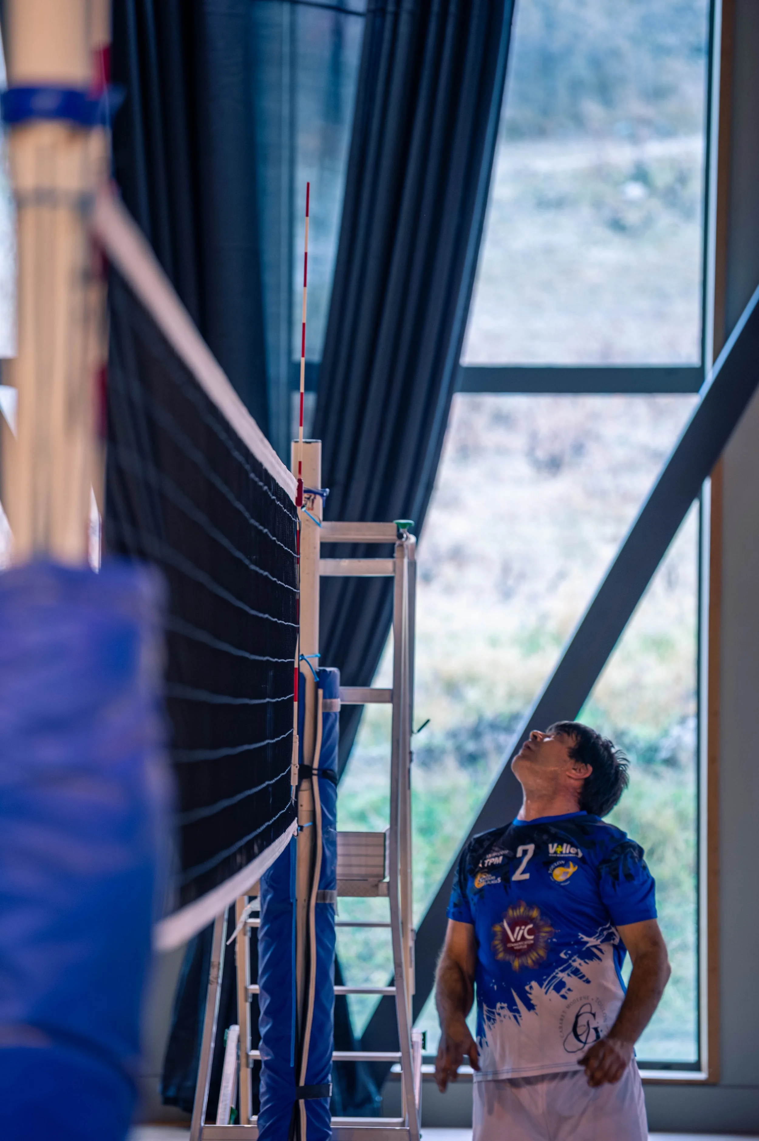Un joueur de volleyball regarde en haut, dans une salle intérieure avec de grandes fenêtres et des rideaux bleus. Il porte un maillot de volley-ball bleu avec le numéro 2.