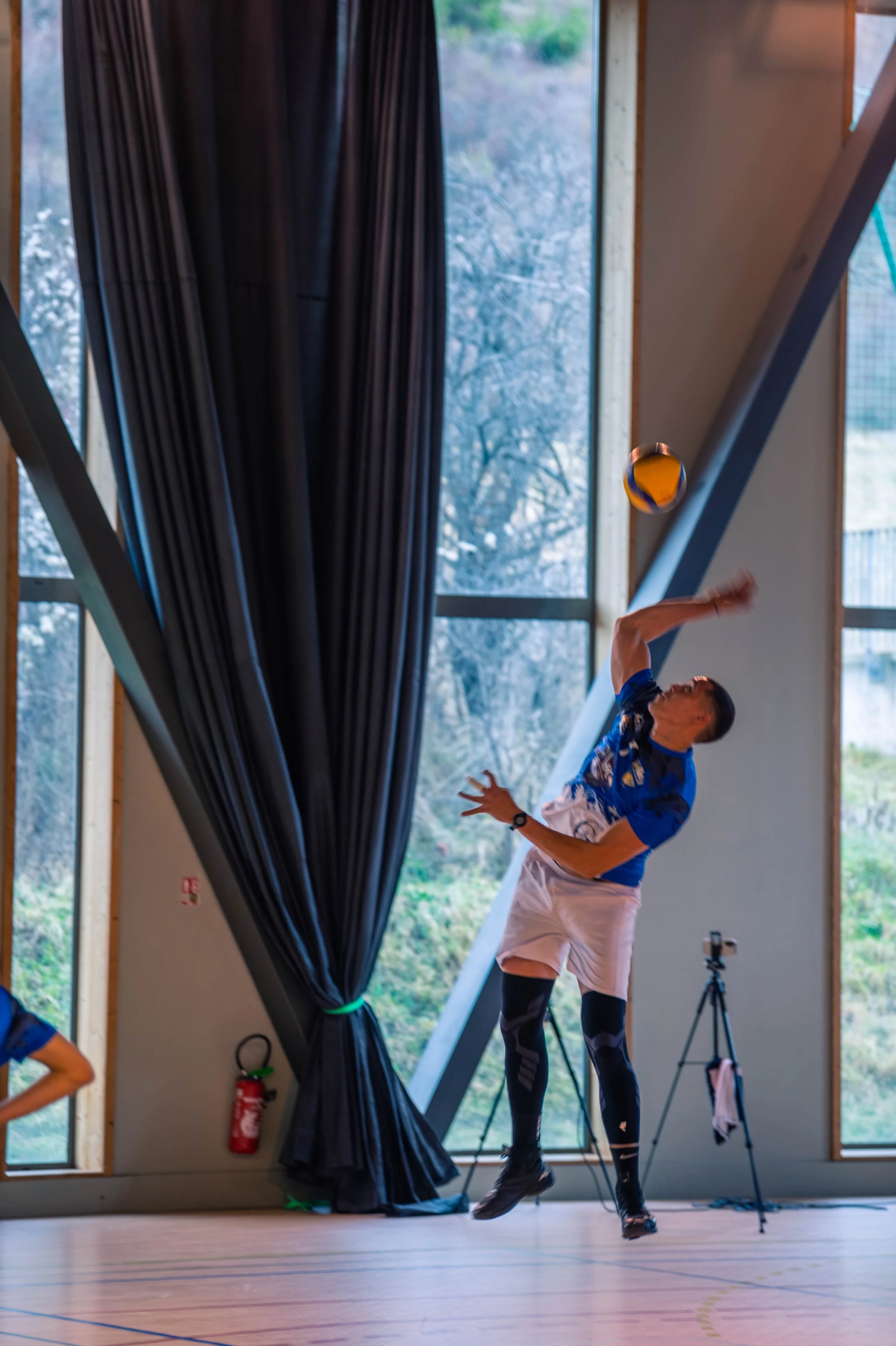 Un homme joue au volleyball en intérieur, il saute pour frapper le ballon, dans une salle avec de grandes fenêtres et des rideaux noirs.