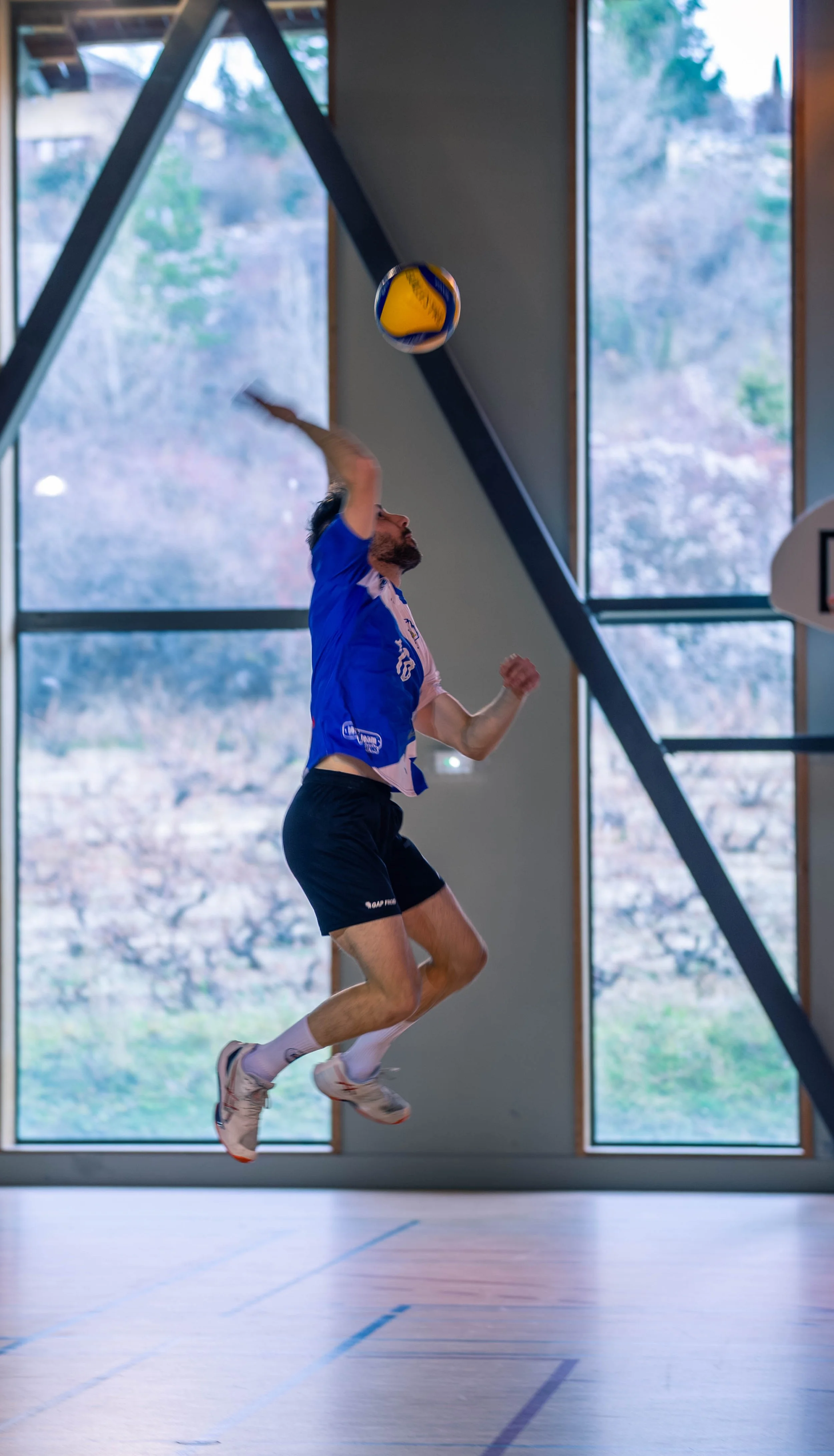 Un joueur de volleyball en train de sauter et de frapper le ballon dans une salle d'intérieur avec de grandes fenêtres derrière lui.