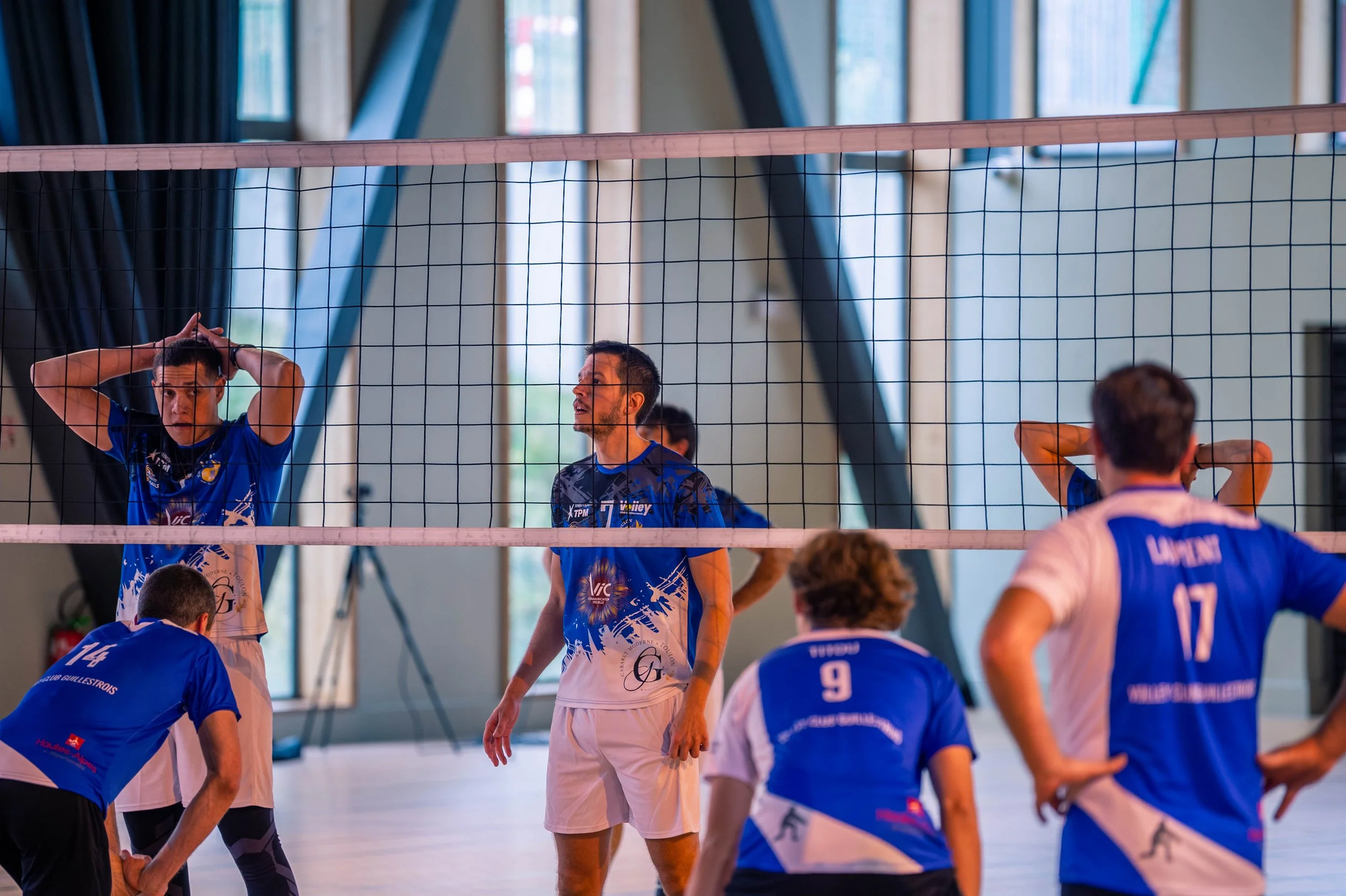 Équipe de volleyball en entraînement en intérieur, joueurs en position, certains avec les mains derrière la tête.