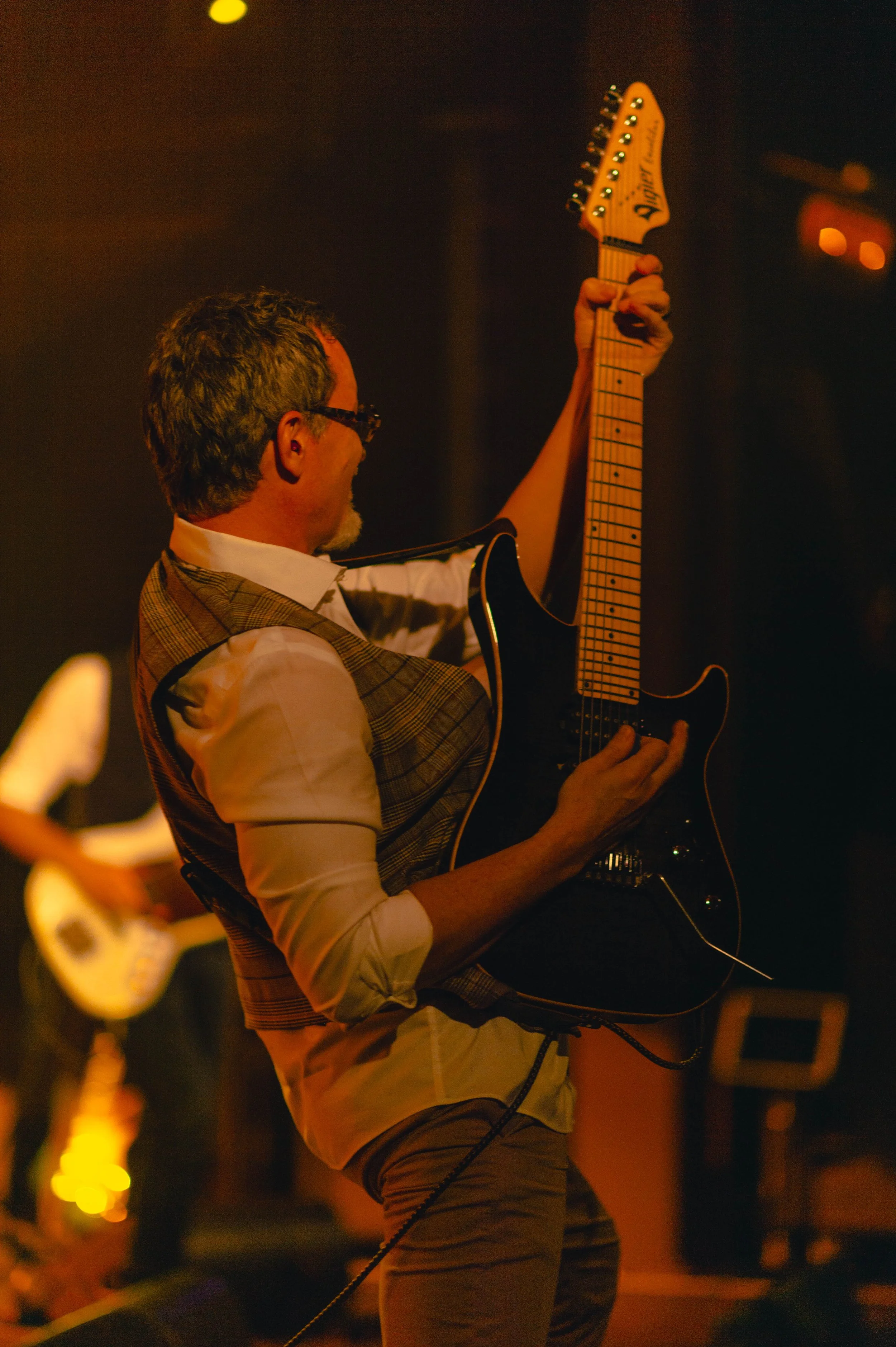 Un homme jouant de la guitare électrique sur scène, éclairé par une lumière chaude, avec une autre personne en arrière-plan jouant également de la guitare.