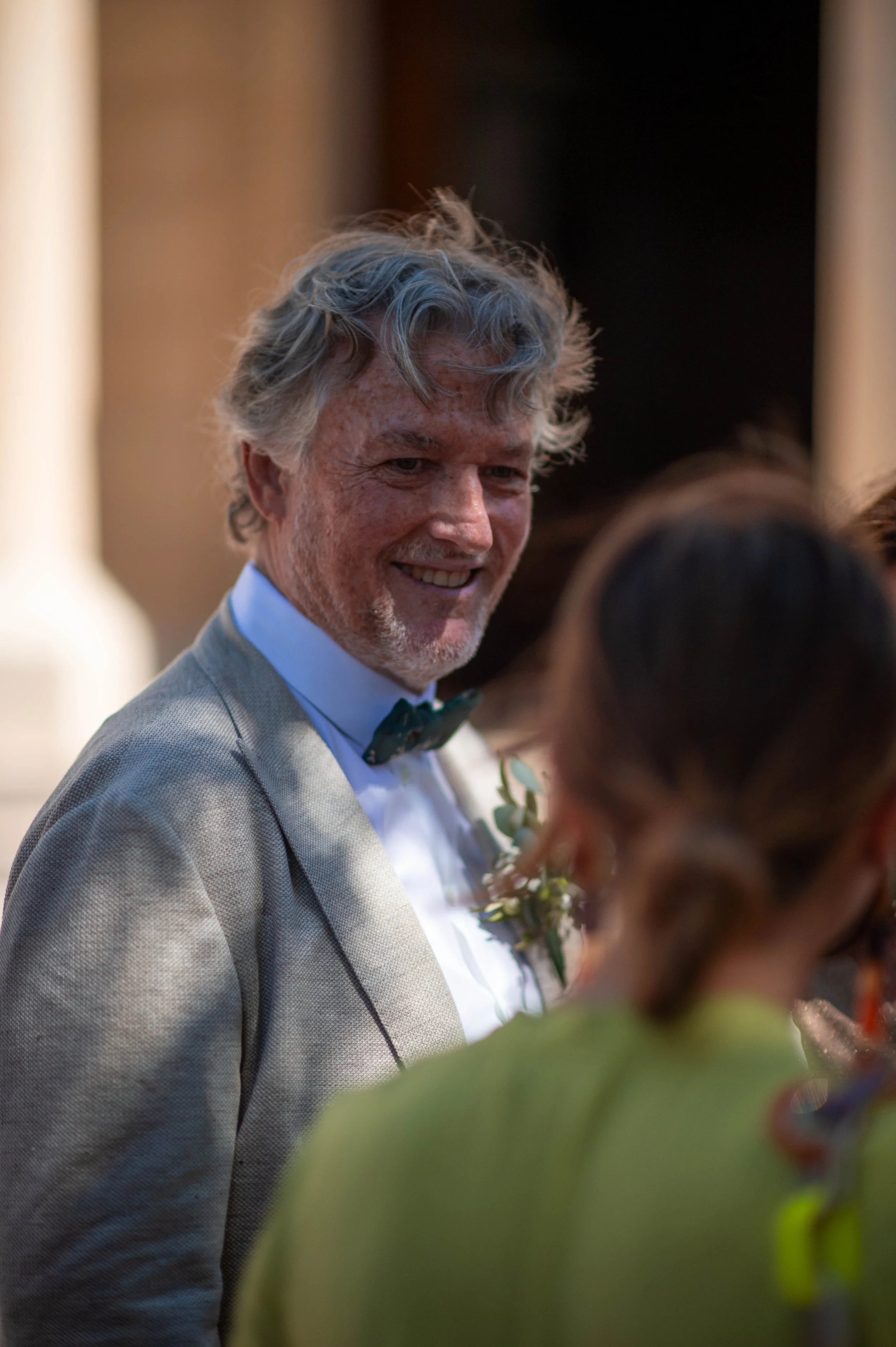 Un homme âgé avec des cheveux gris, portant un costume beige, une chemise blanche et un nœud papillon, sourit lors d'un événement social ou un mariage.