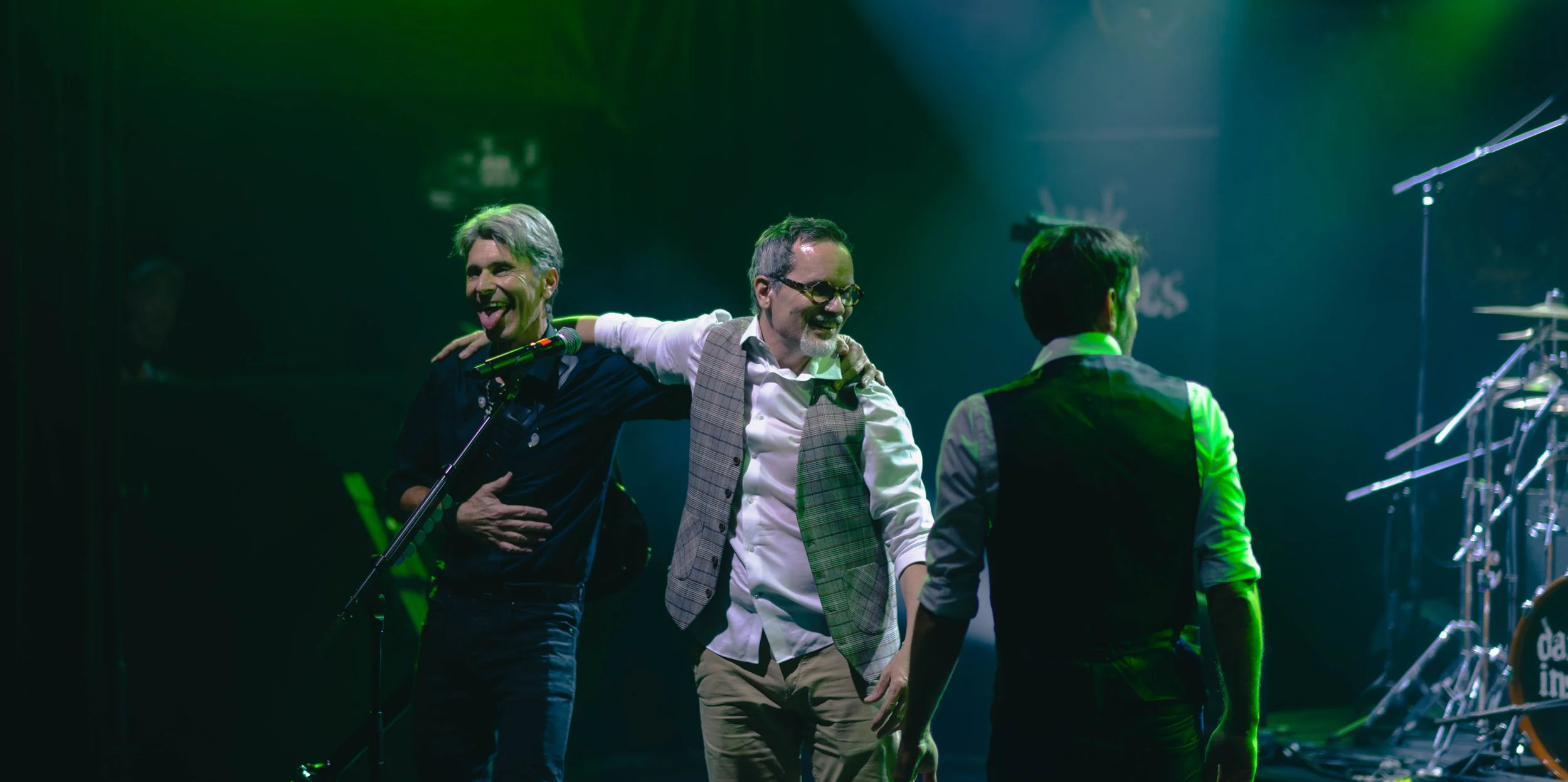 Trois hommes sur scène, l'air heureux, en train de prendre un moment de convivialité lors d'un concert, avec un fond lumineux vert et des instruments de musique.