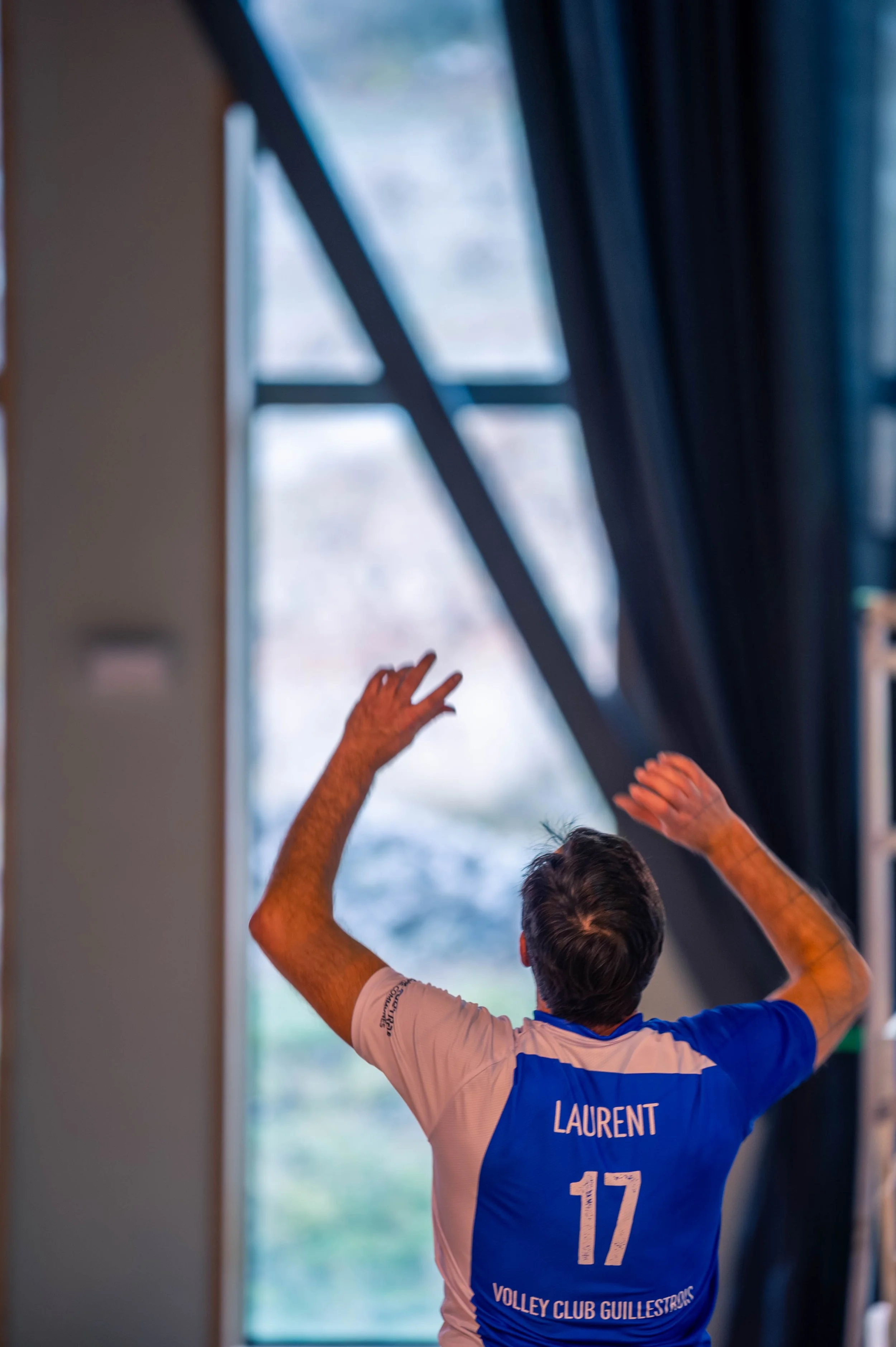 Un homme vu de dos, portant un maillot de volley avec le nom 'LAURENT' et le numéro 17, levant les bras dans une salle avec de grandes fenêtres et des rideaux bleus.
