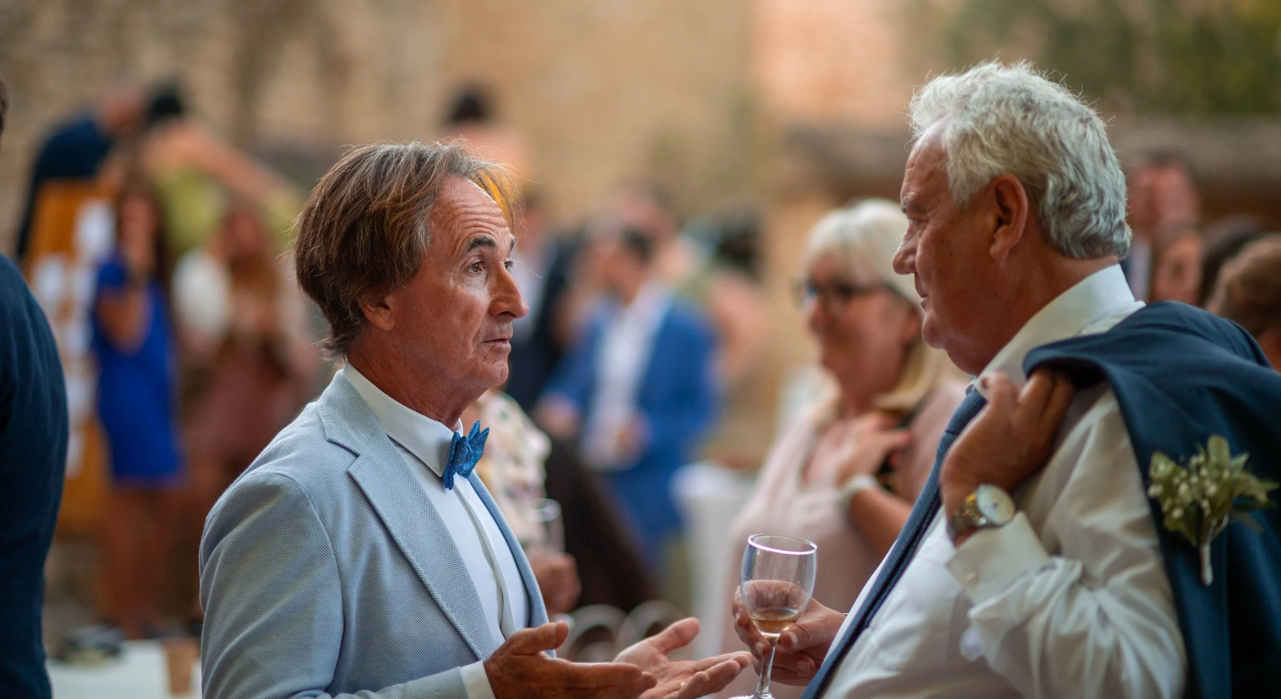 Deux hommes en tenue formelle discutent à une fête, un d'eux tient un verre de vin et porte une veste sur l'épaule.