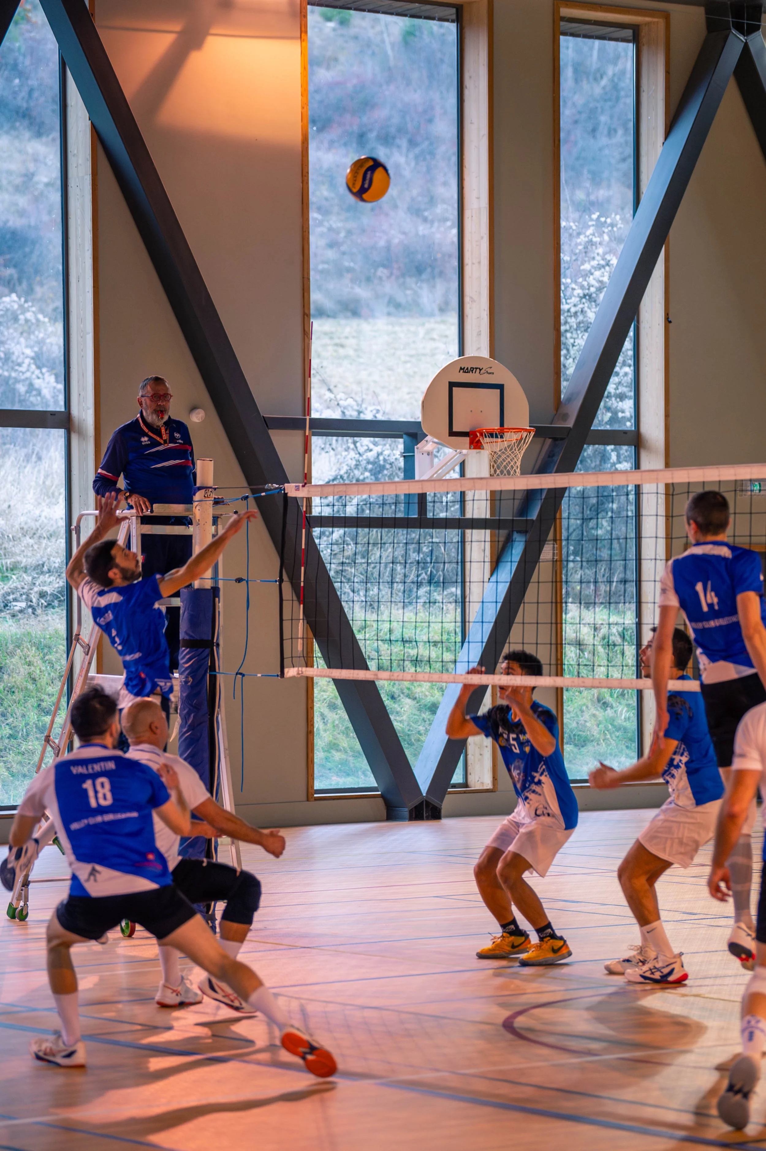 Une équipe de volley-ball en action à l'intérieur d'un gymnase avec un ballon suspendu dans l'air et un joueur prêt à sauter près du filet, un arbitre à gauche, et un panier de basketball fixé au mur derrière la vitre.