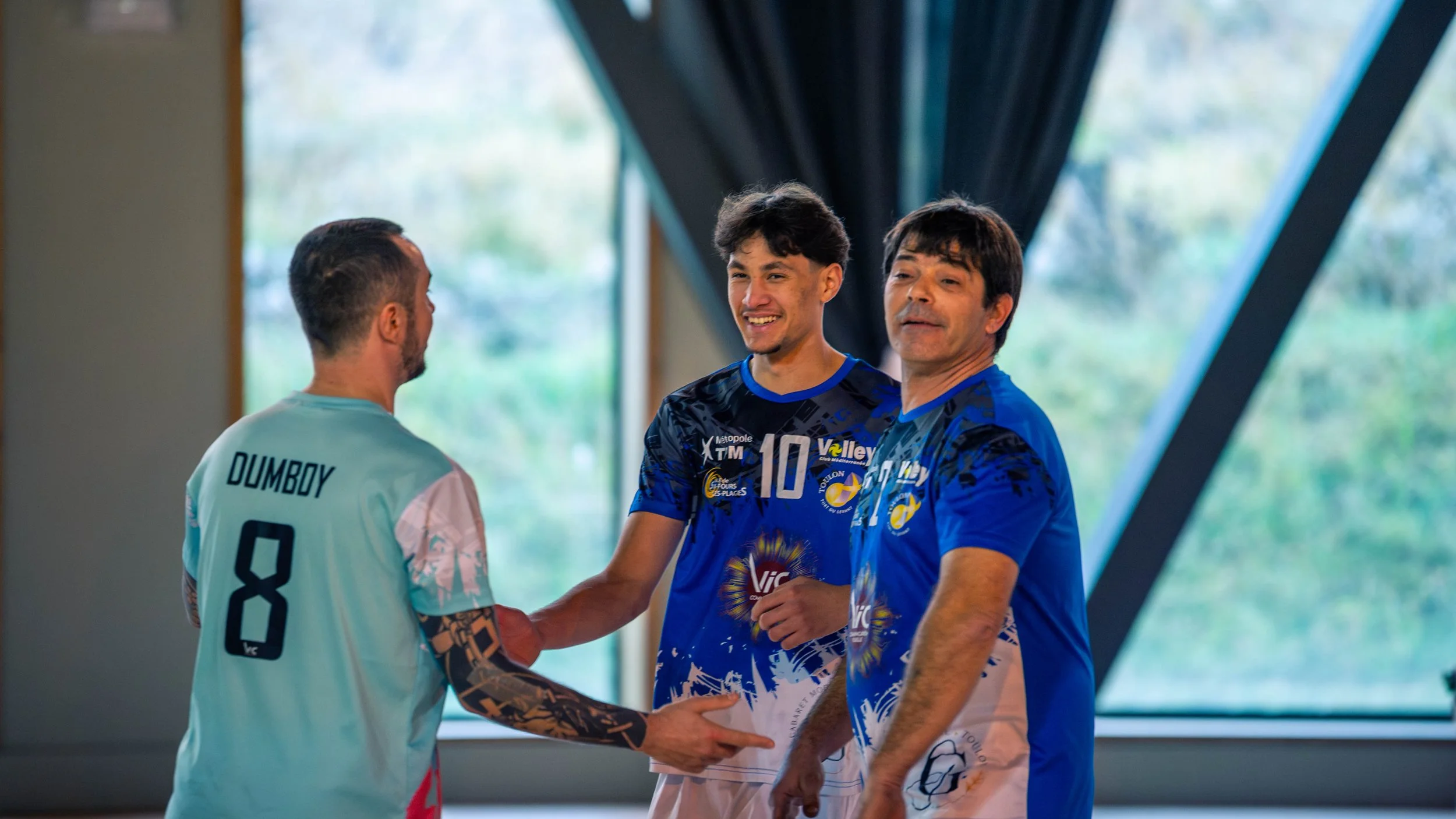 Trois hommes dans un gymnase, deux portent des maillots de volleyball, l'un d'eux serre la main du troisième.
