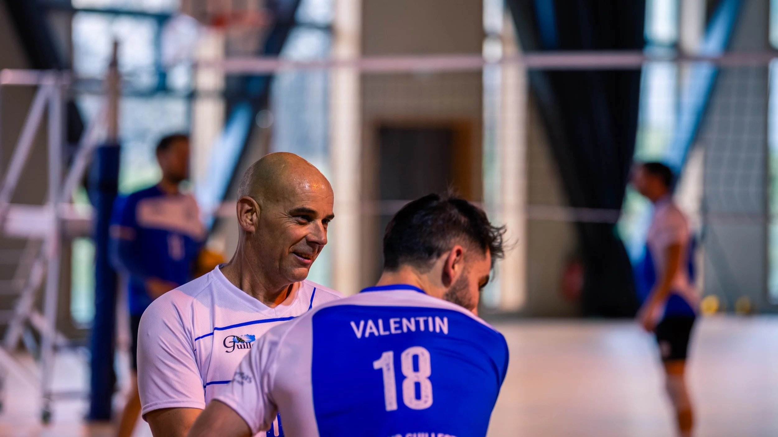 Deux joueurs de volley-ball en tenues bleues et blanches, avec un nom et un numéro, discutent dans une salle de sport intérieure. Deux autres personnes sont visibles en arrière-plan, une dans une tenue bleue et blanche, l'autre en blanc avec des déta