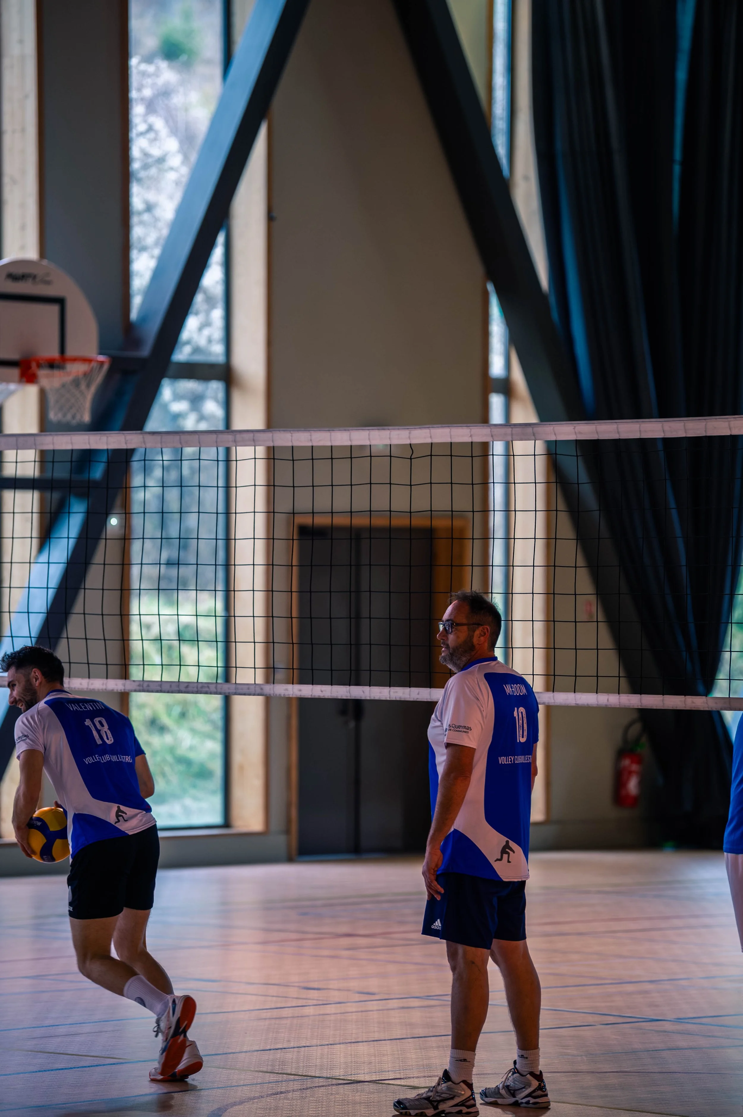 Deux hommes jouent au volleyball en intérieur, avec un filet de volleyball, un ballon, et un décor d'architecture moderne avec grandes fenêtres.