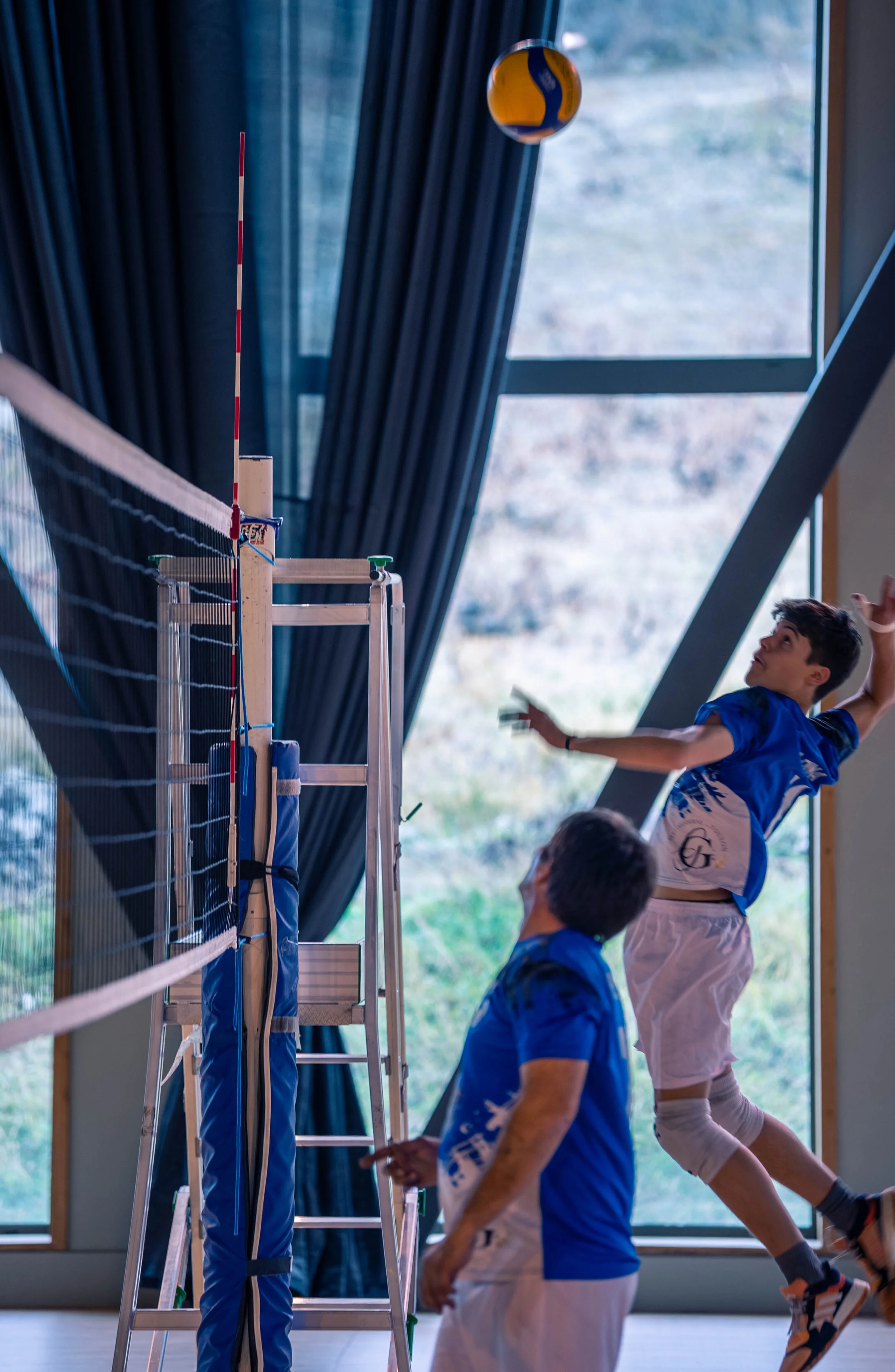 Deux jeunes jouant au volley-ball en intérieur, l'un sautant pour frapper le ballon et l'autre observant, avec un filet de volleyball et une grande fenêtre en arrière-plan.
