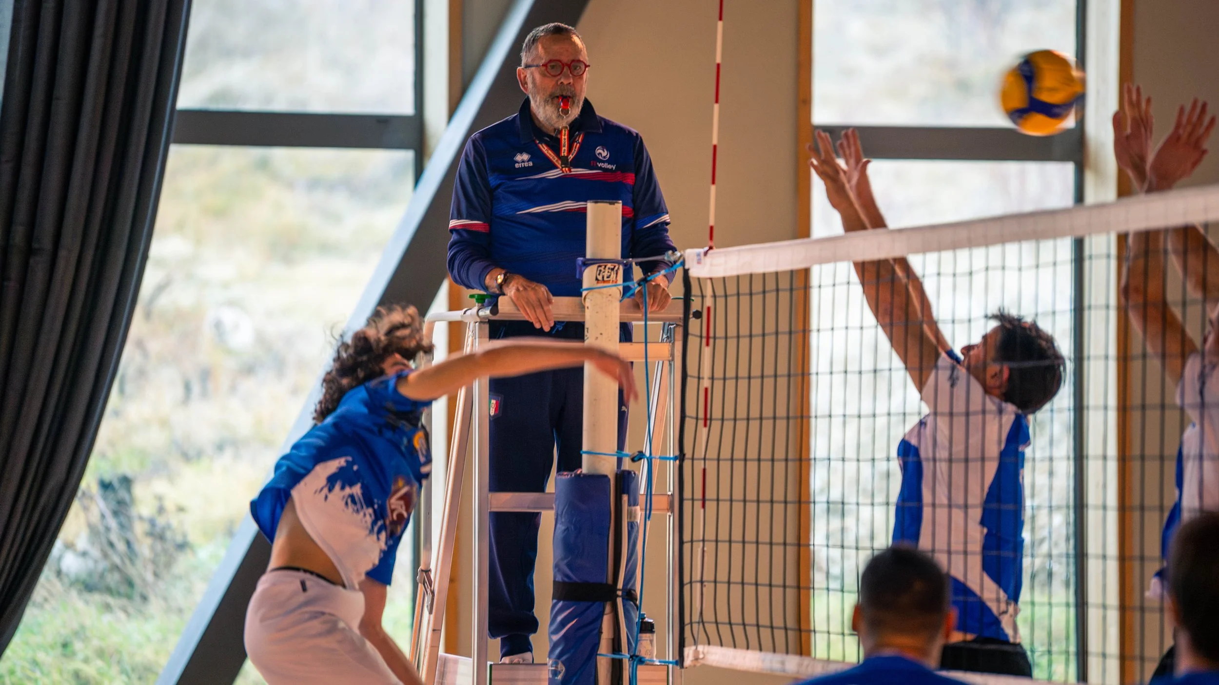 Entraîneur de volleyball en train de donner des instructions lors d'un match dans une salle en intérieur, avec des joueurs tentant de bloquer un ballon.