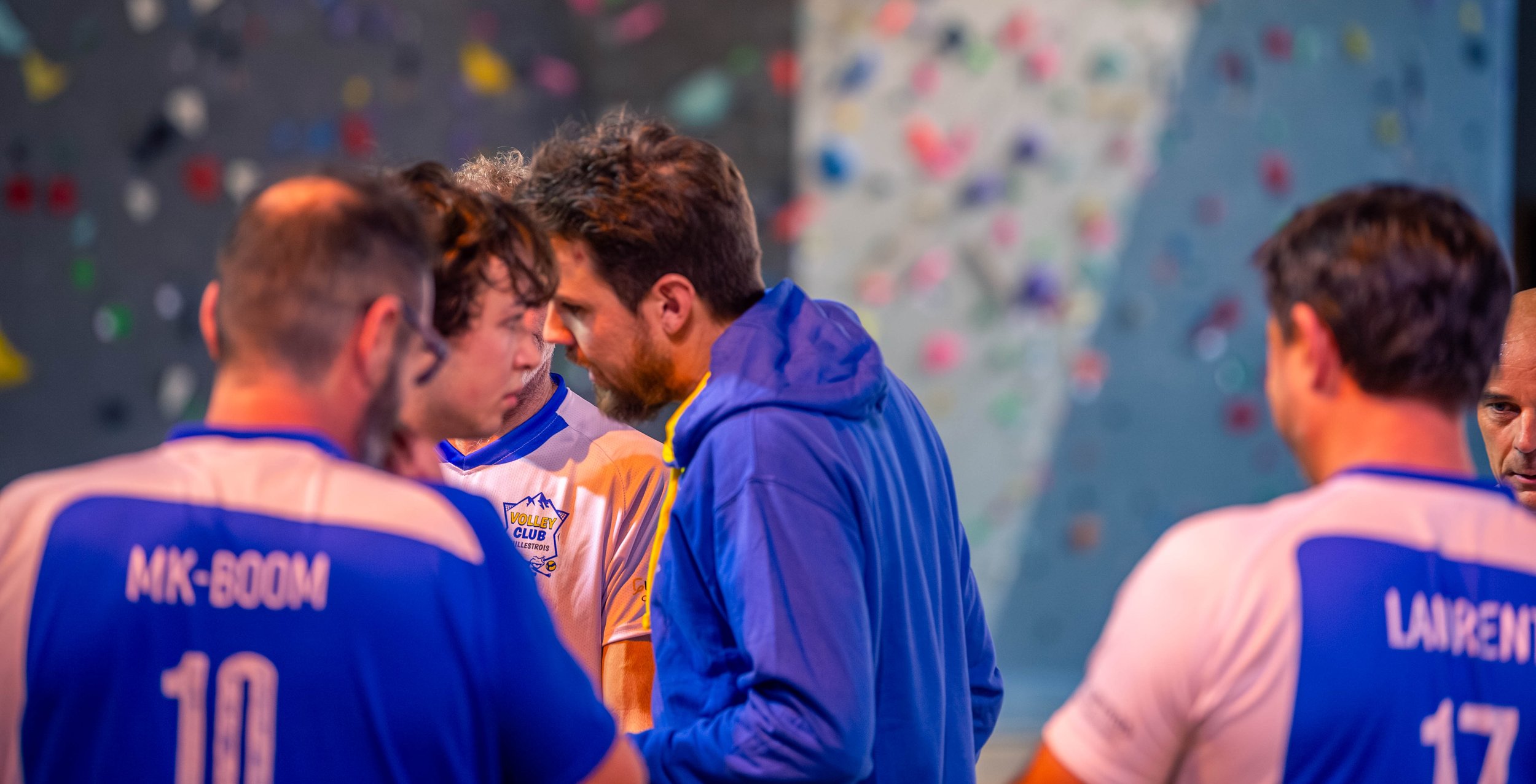 Groupe de joueurs de volleyball en discussion avec un entraîneur ou un coéquipier dans une salle d'escalade ou de sport, avec un mur d'escalade en fond.