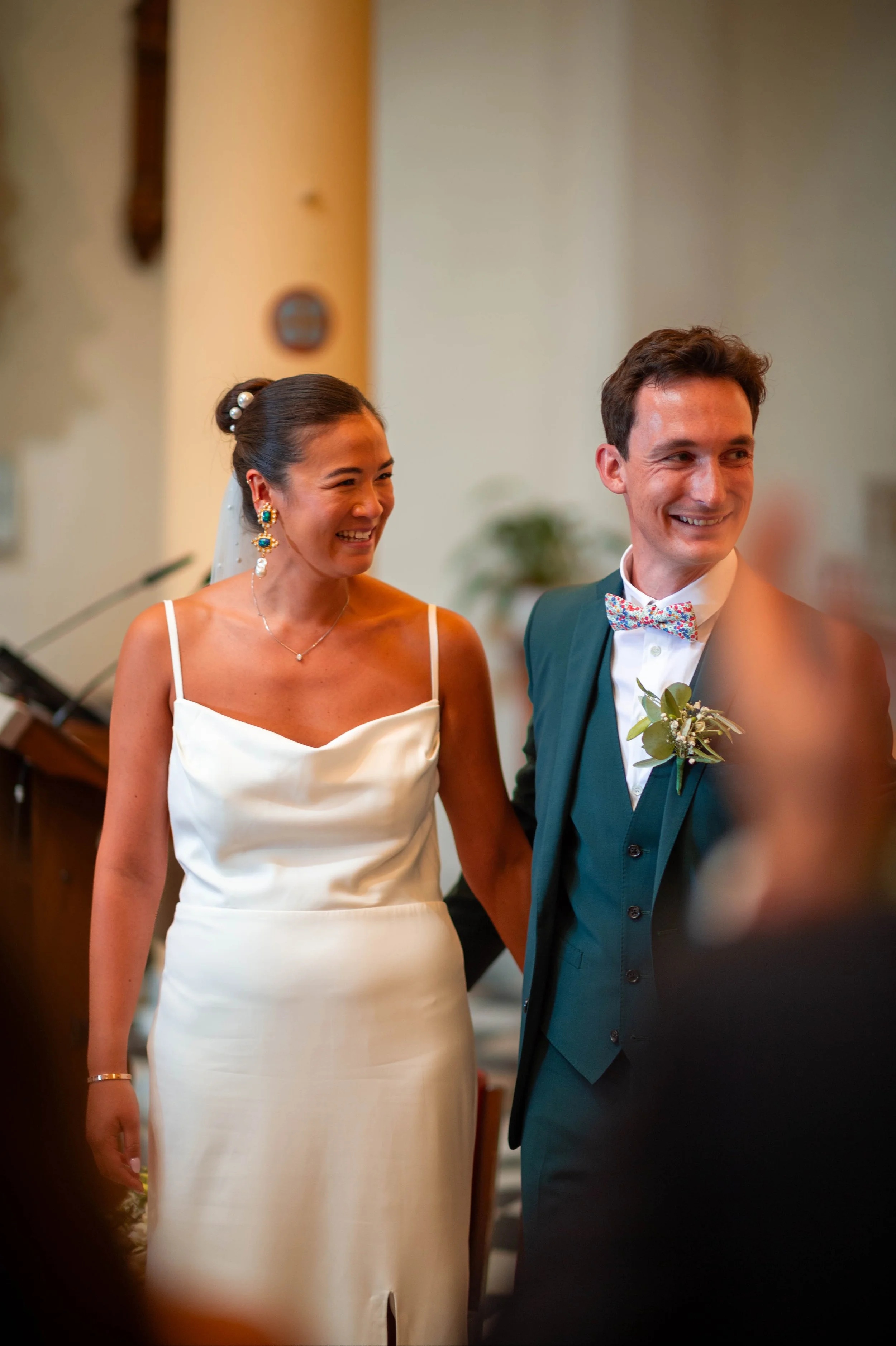 Un couple de mariés souriants lors de leur mariage, la mariée porte une robe blanche à bretelles et la mariée un costume vert avec un nœud papillon coloré.